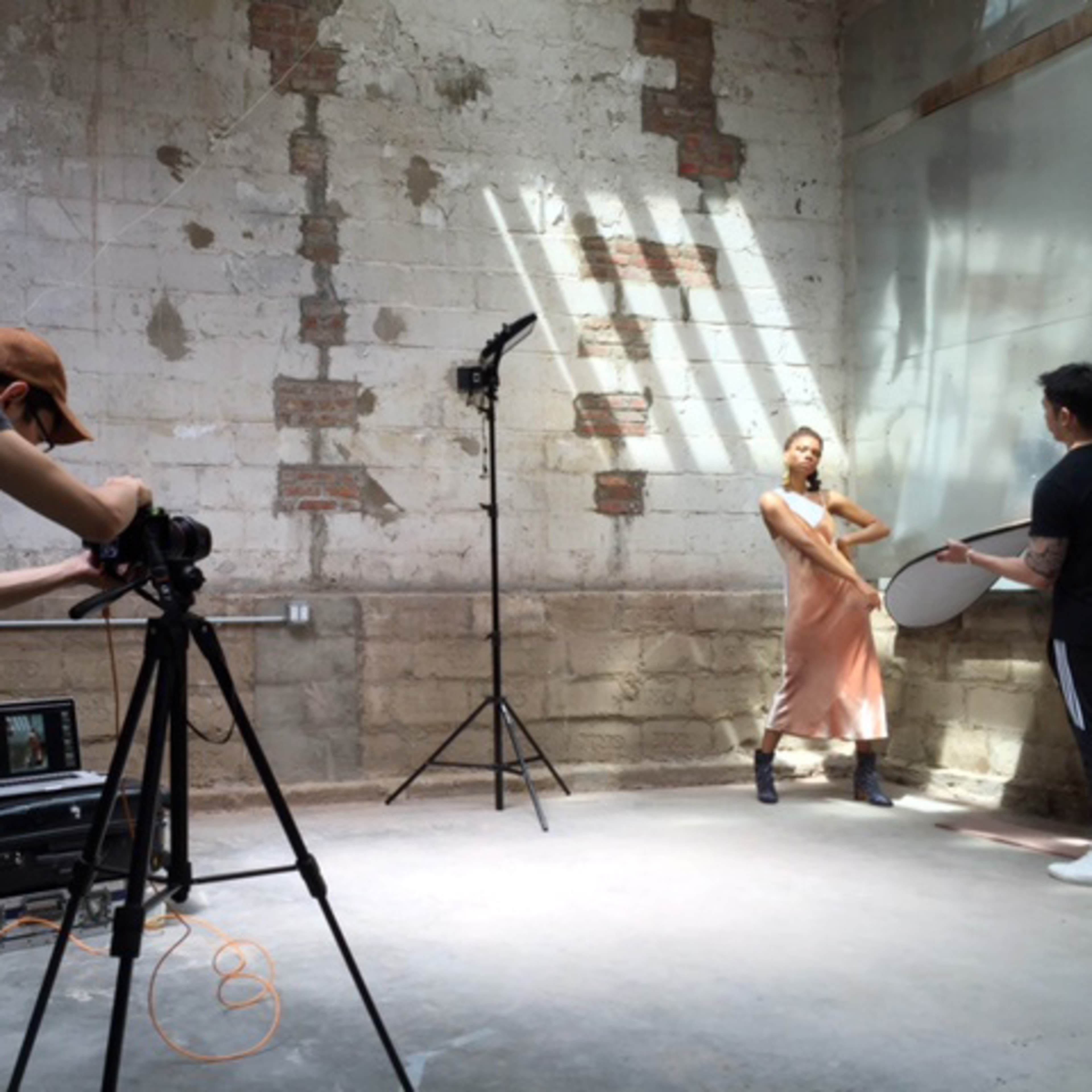 A photographer captures a model posing in a studio with exposed brick walls while a team sets up lighting and reflects light with a silver disc.