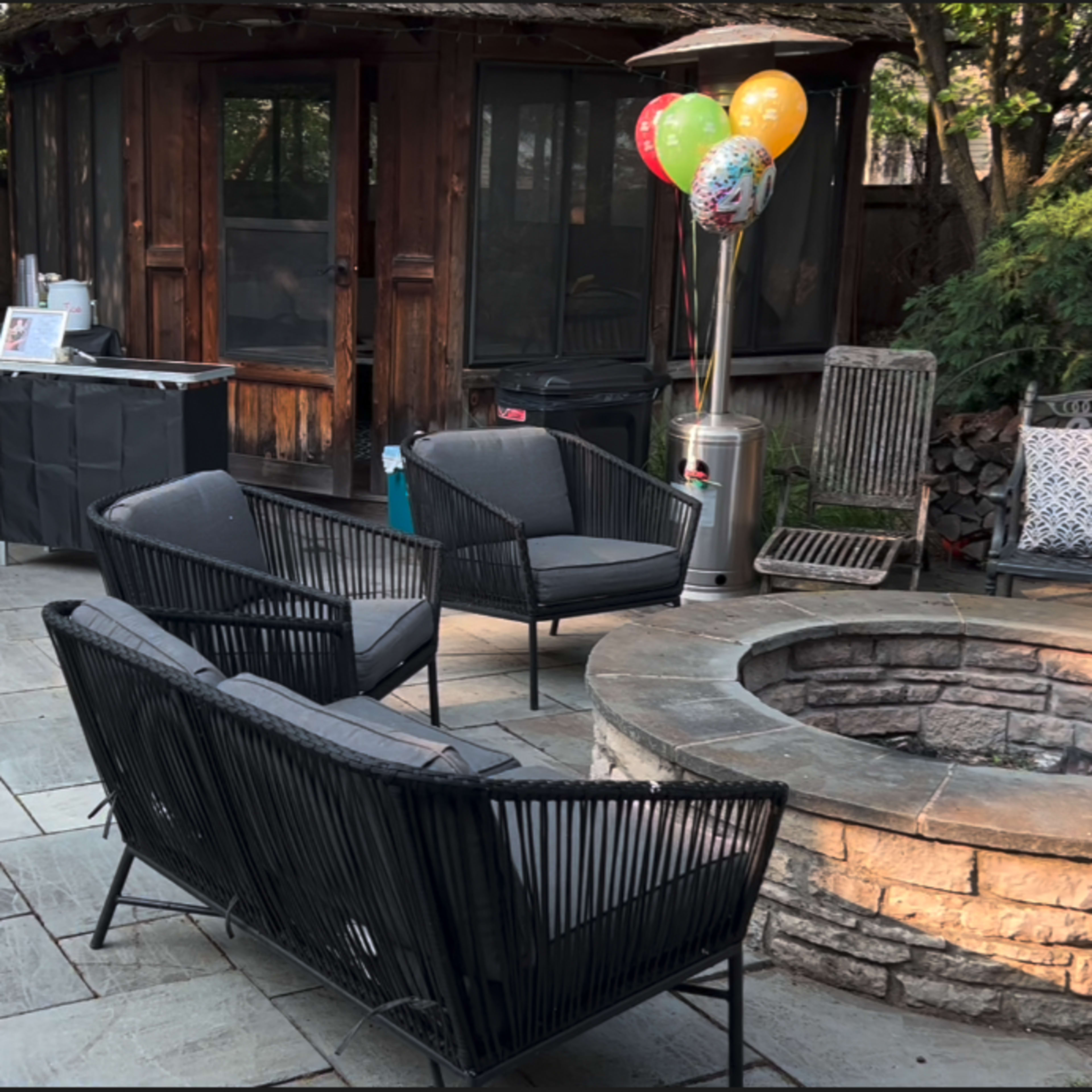 The patio features a stone fire pit surrounded by black wicker chairs, a grill, and balloons tied to a post.