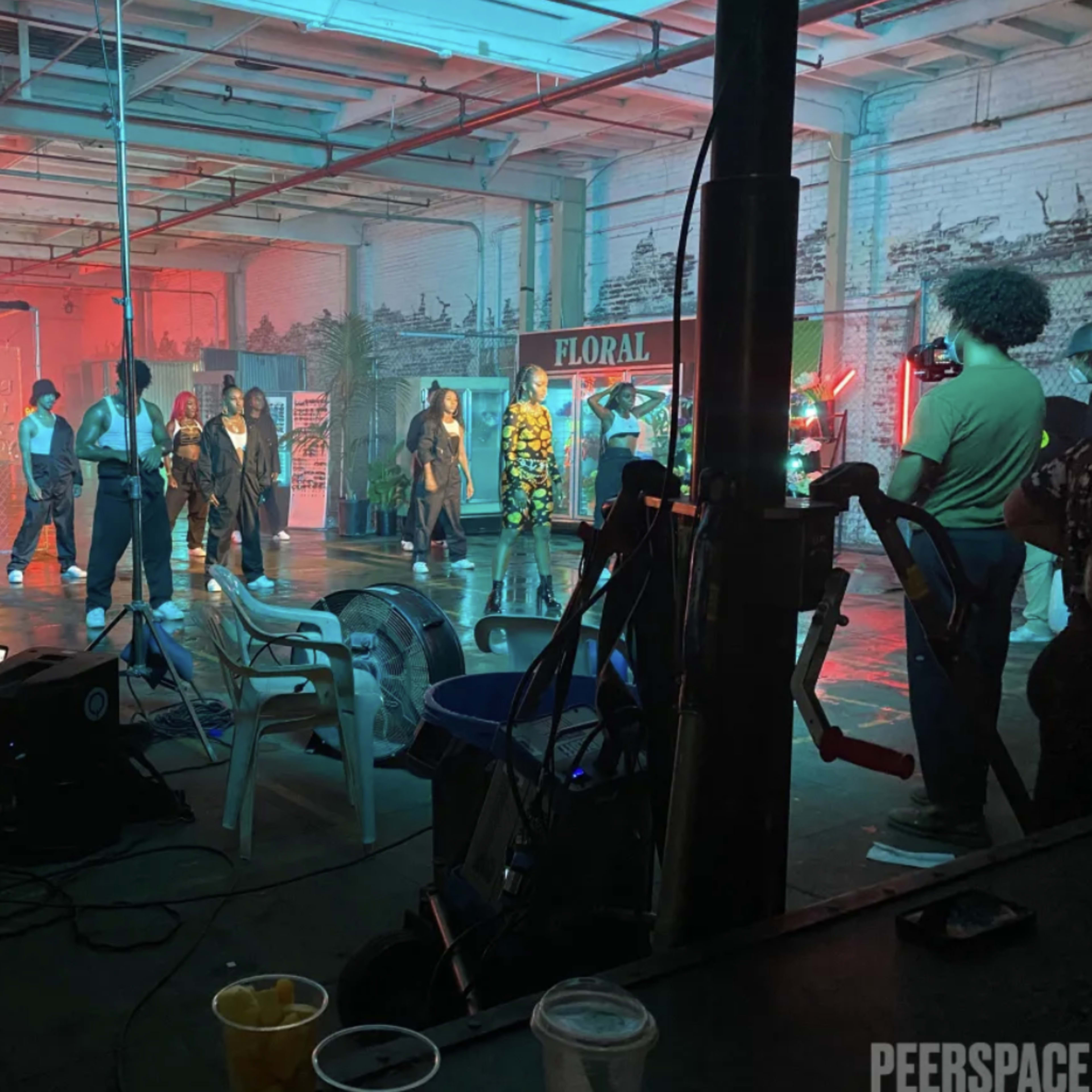 A group of performers is rehearsing on a brightly lit stage set in an industrial space, while crew members operate equipment nearby.
