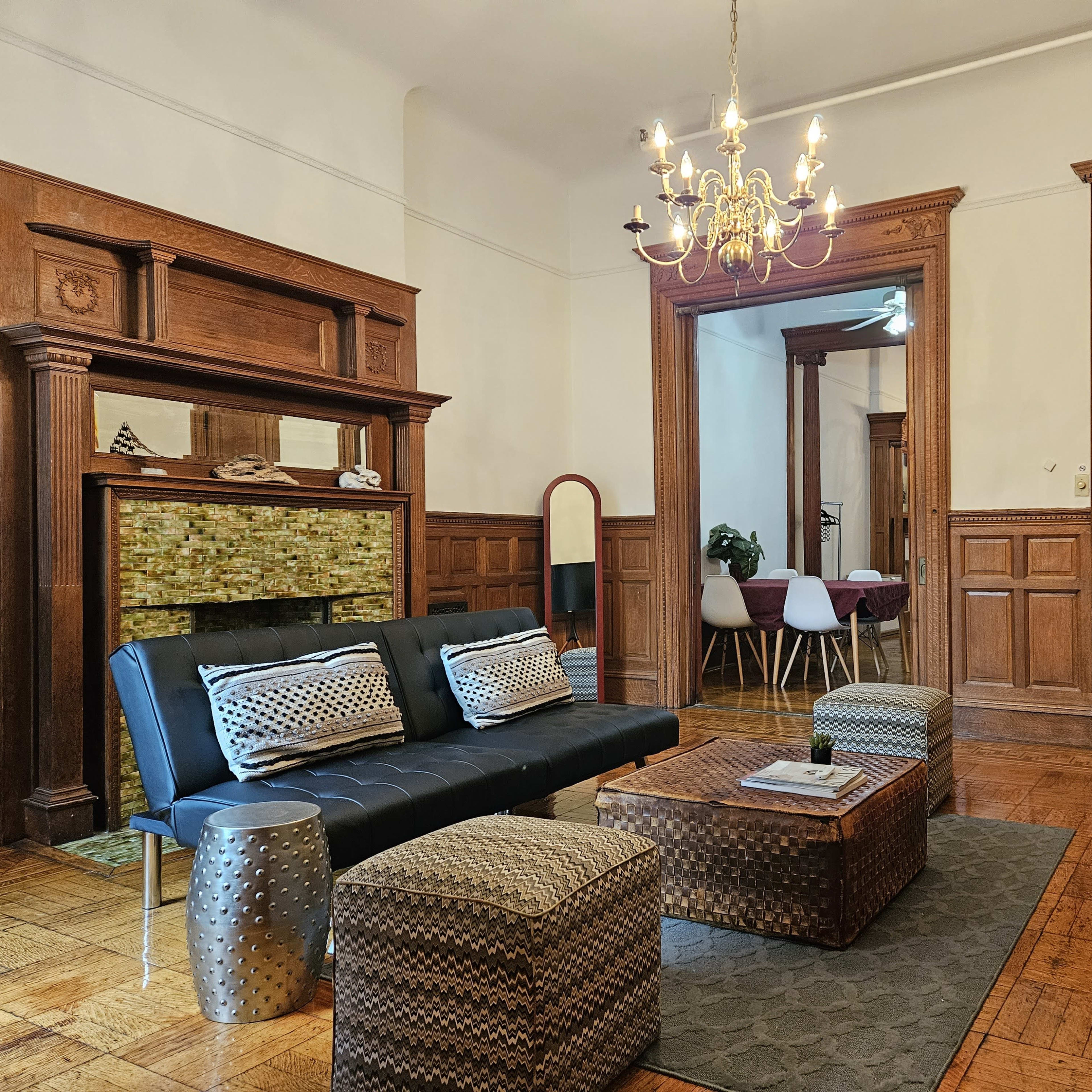 A living room features a black sofa and a wooden coffee table, surrounded by decorative stools and a chandelier, with paneled walls and a stylish fireplace.