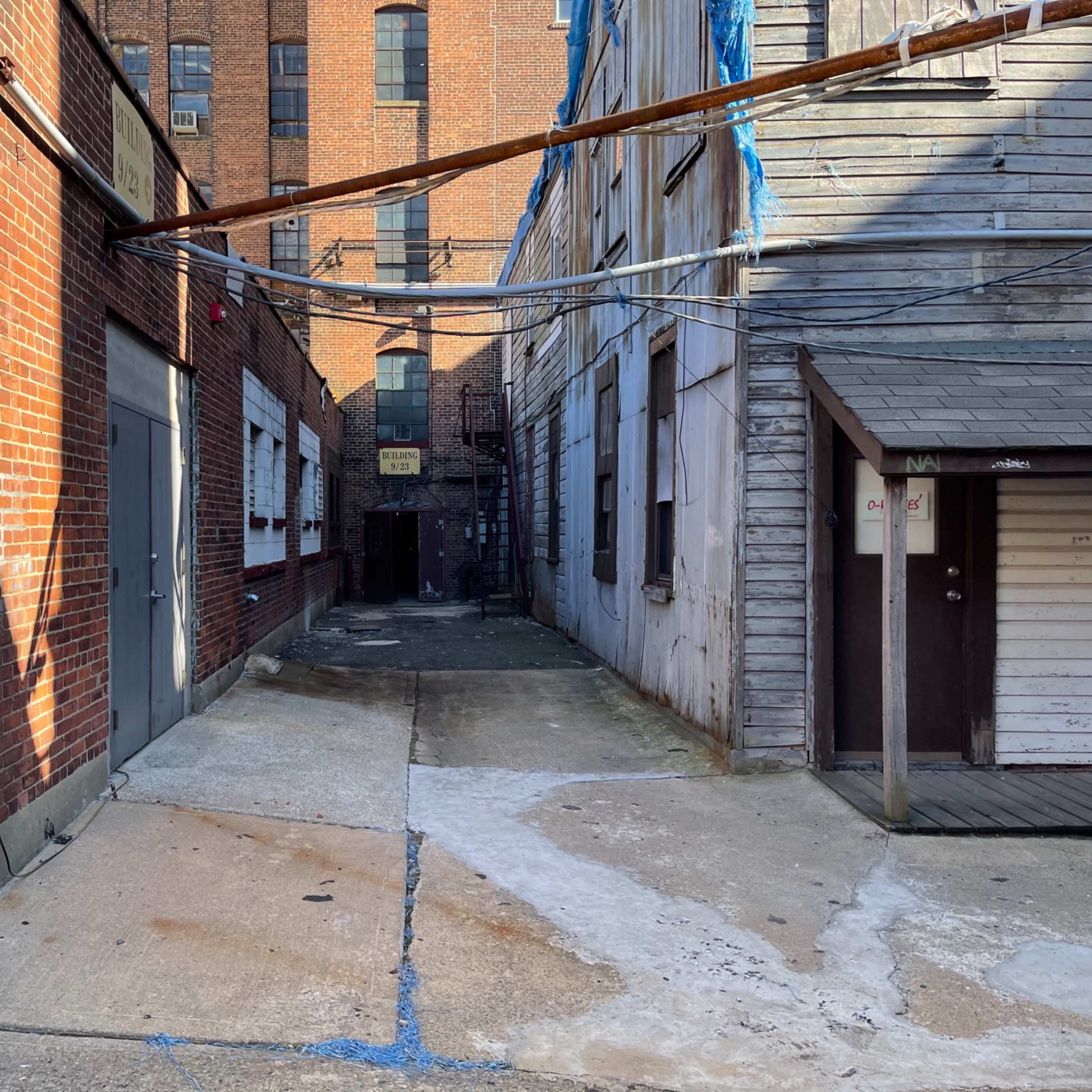 A narrow alleyway between brick and wooden buildings, with concrete pavement and overhead wires.
