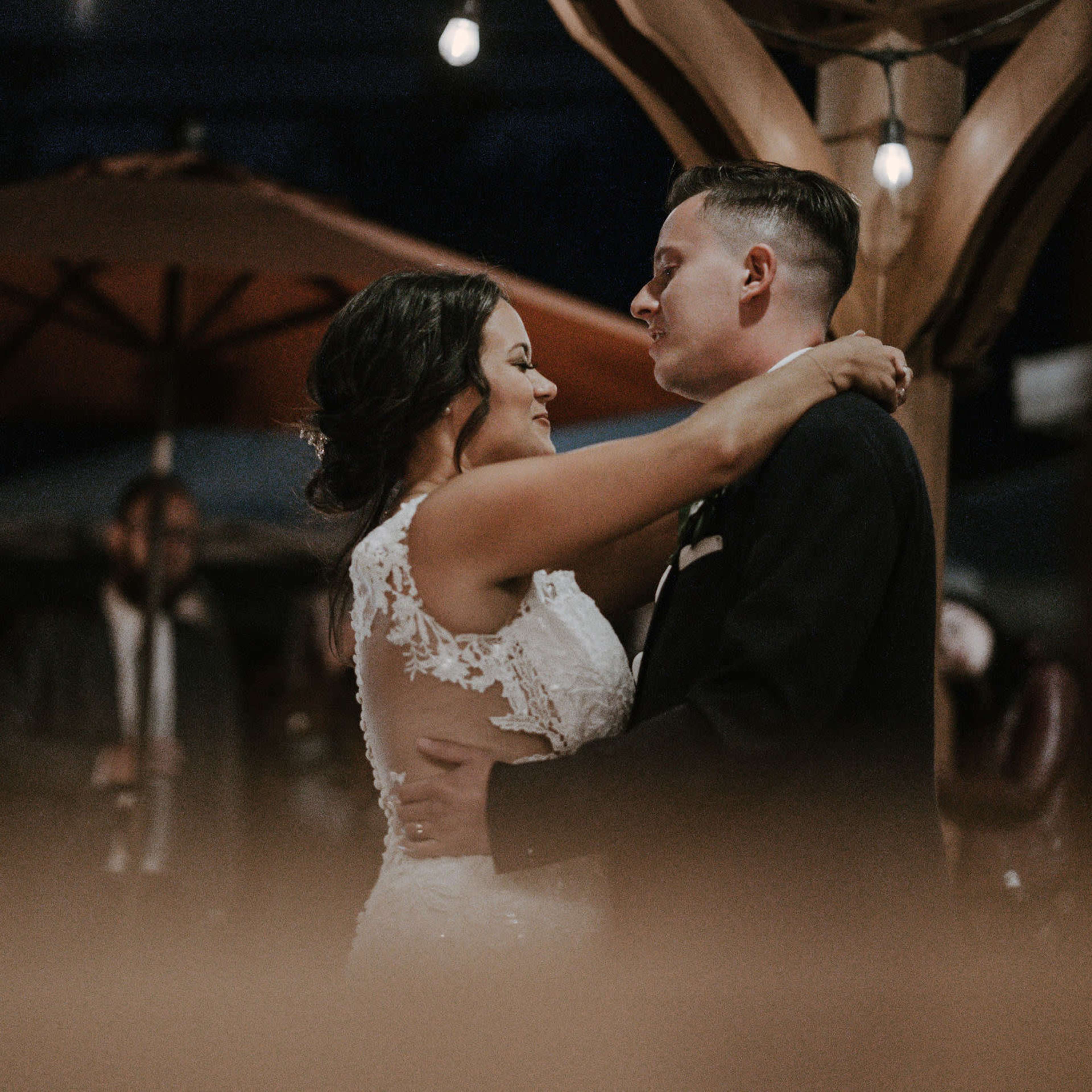 A couple dances closely under dim lights at an outdoor venue, surrounded by guests.