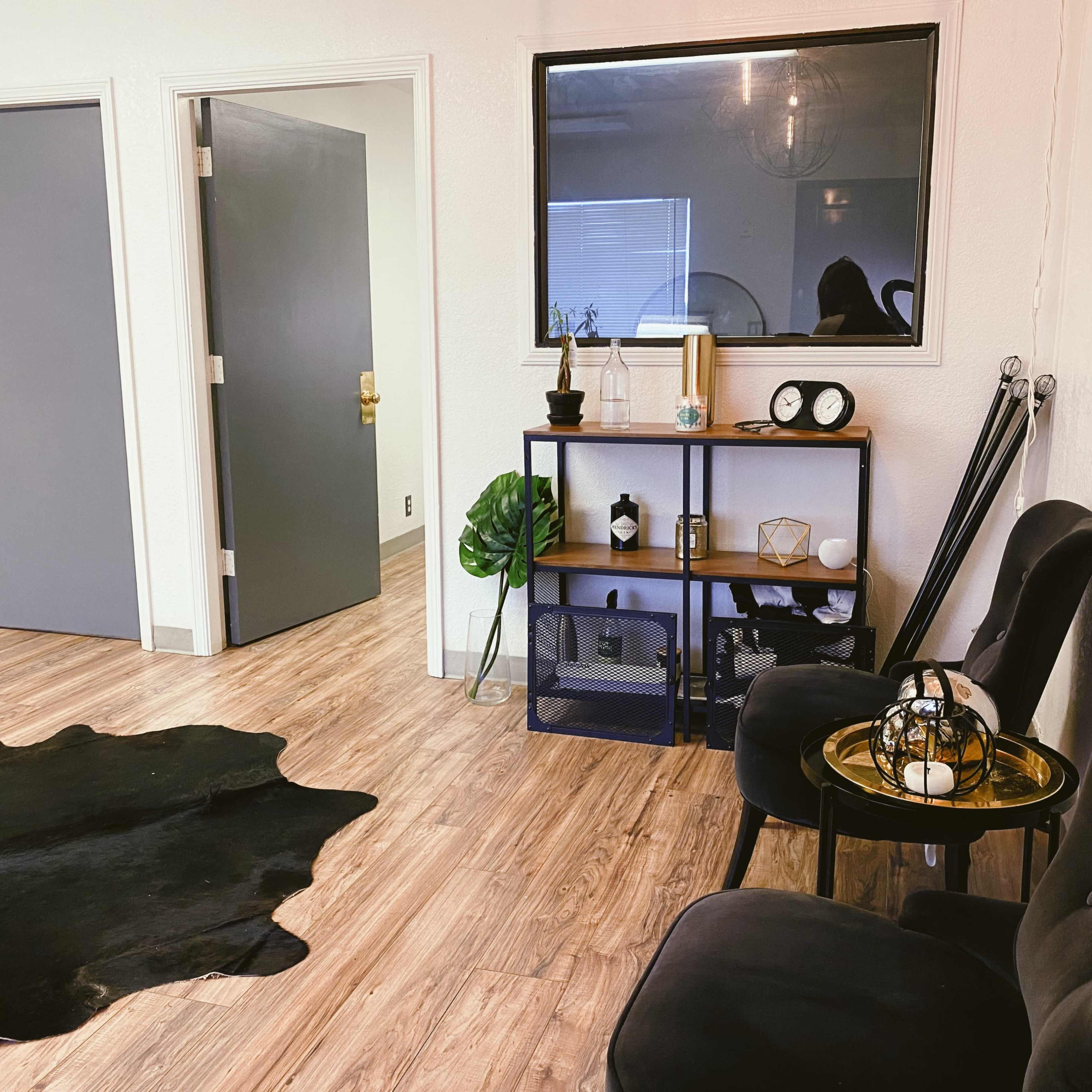 A minimalist interior space featuring a shelf with decor items, a pair of black chairs, a door leading to another room, and a rug on a wooden floor.