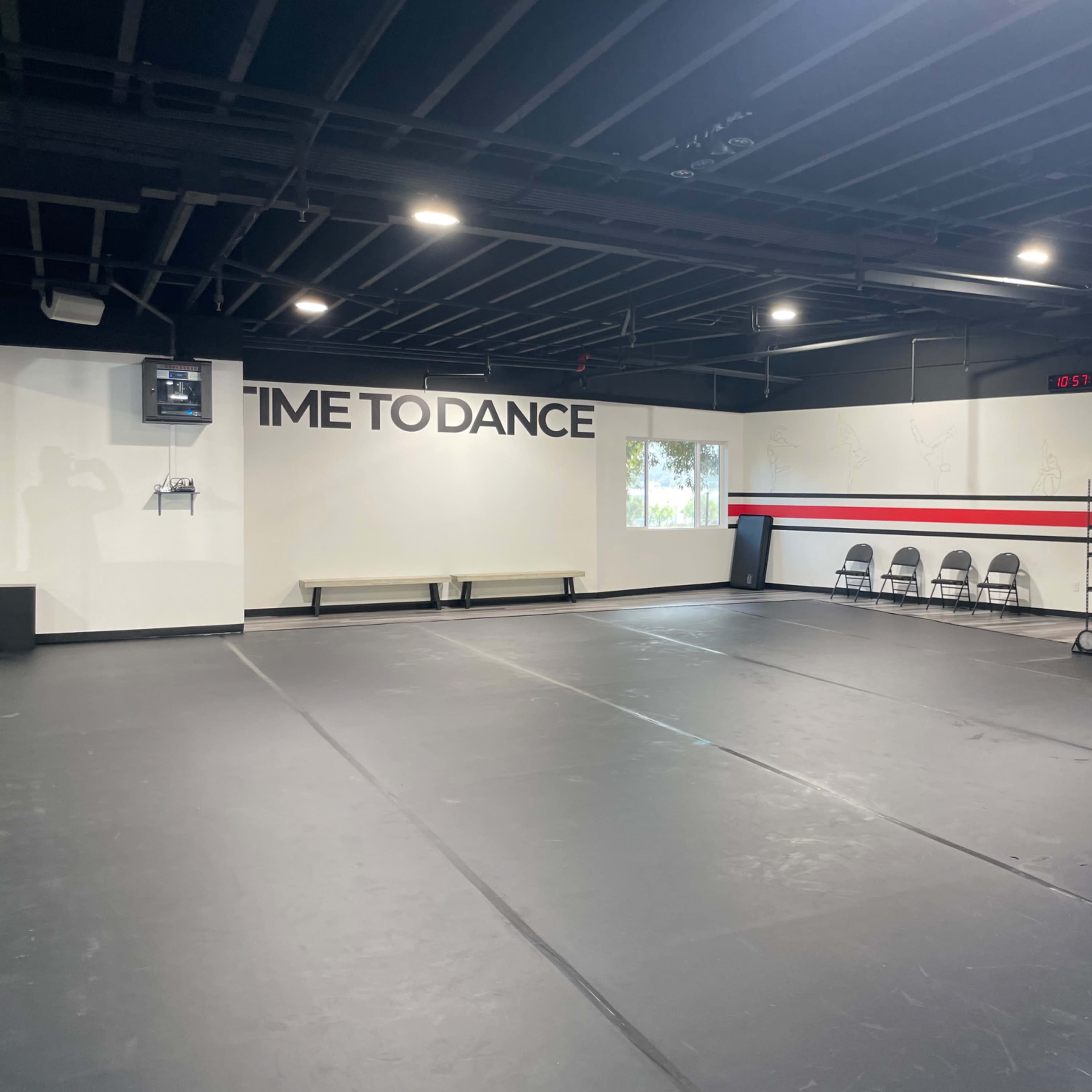 An empty dance studio with a black floor, white walls featuring a "TIME TO DANCE" sign, and a row of chairs along one side.