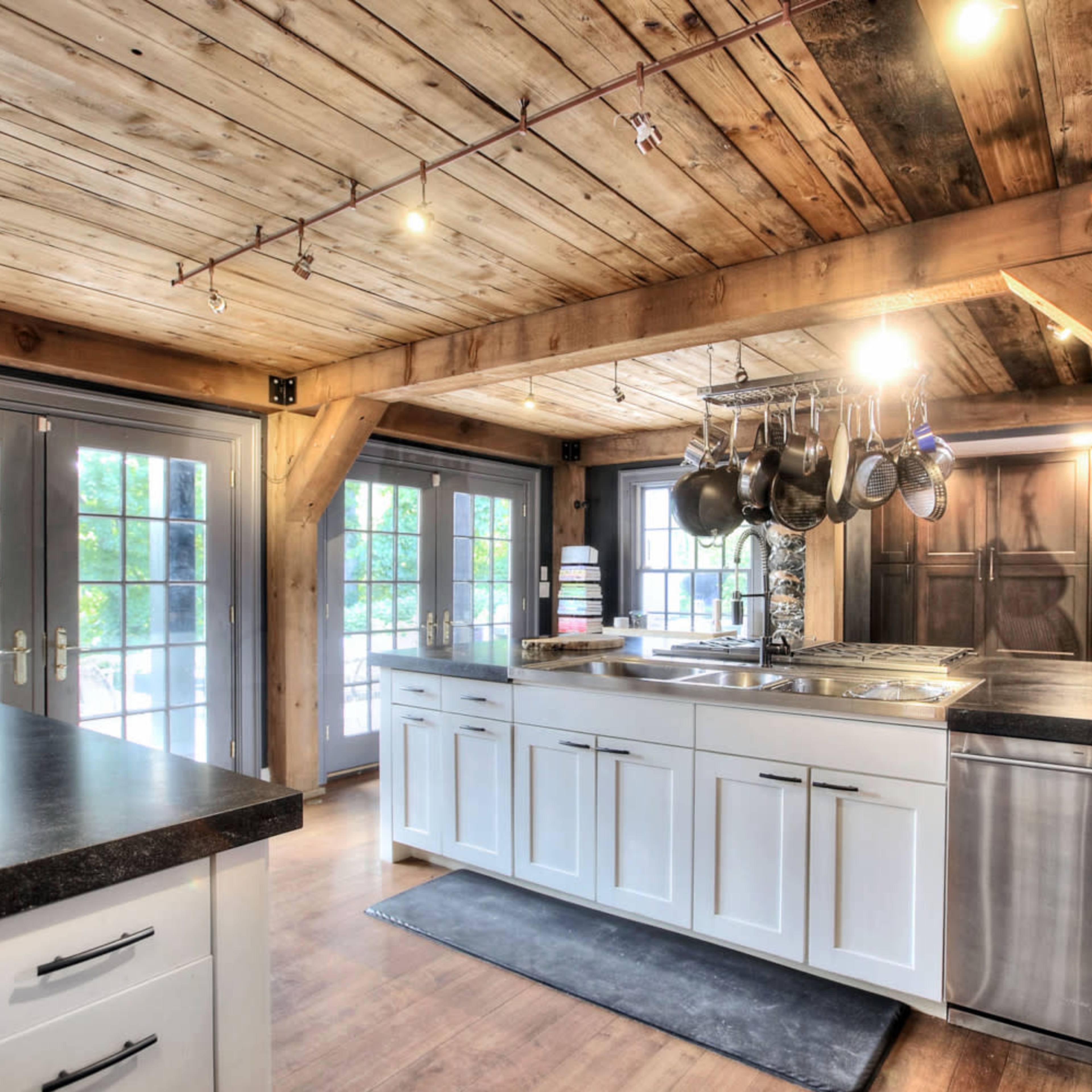 A kitchen with wooden beams and a countertop, featuring pots hanging from a rack above the stove and large windows that provide natural light.