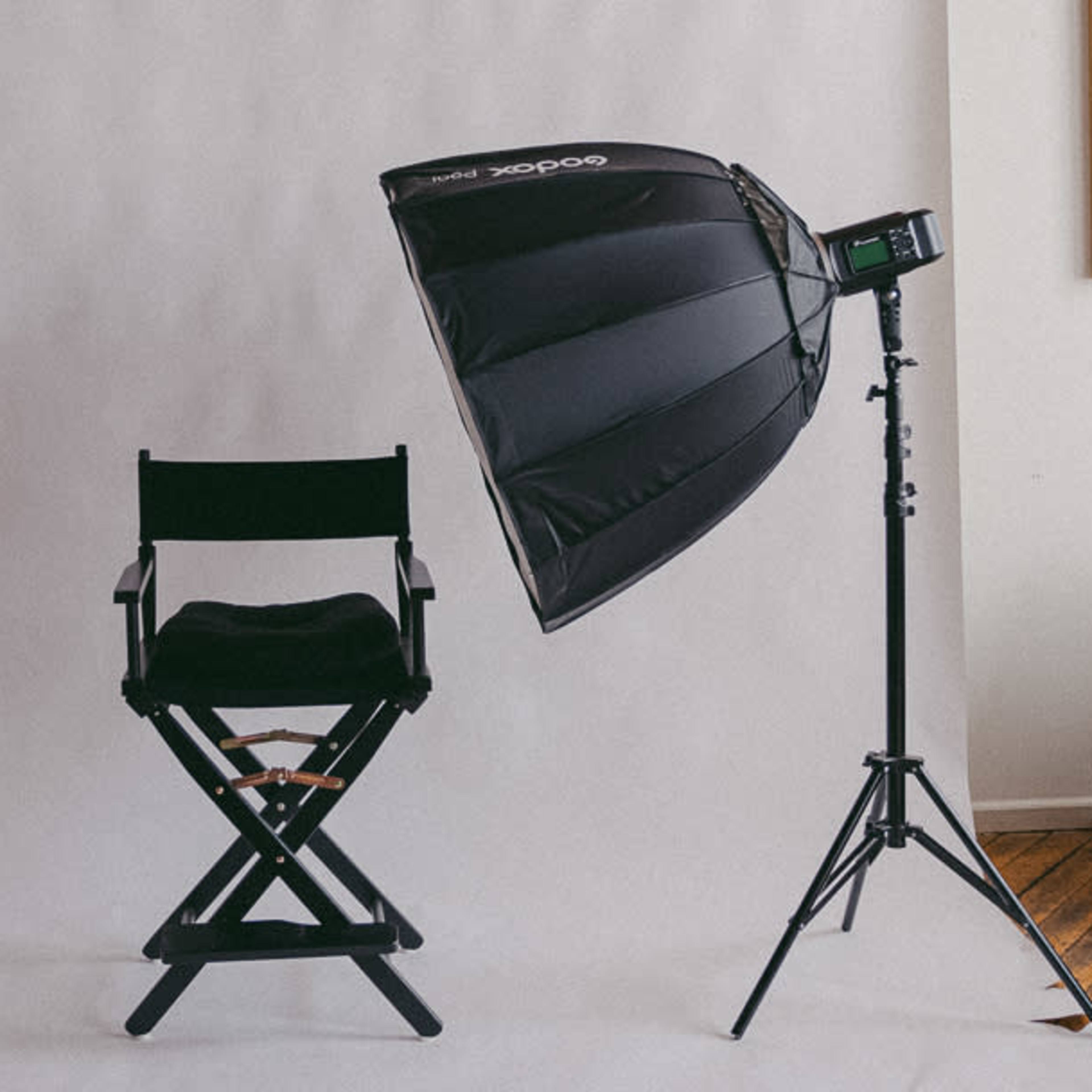 A photography setup featuring a black director's chair beside a light modifier mounted on a stand, all against a white backdrop.