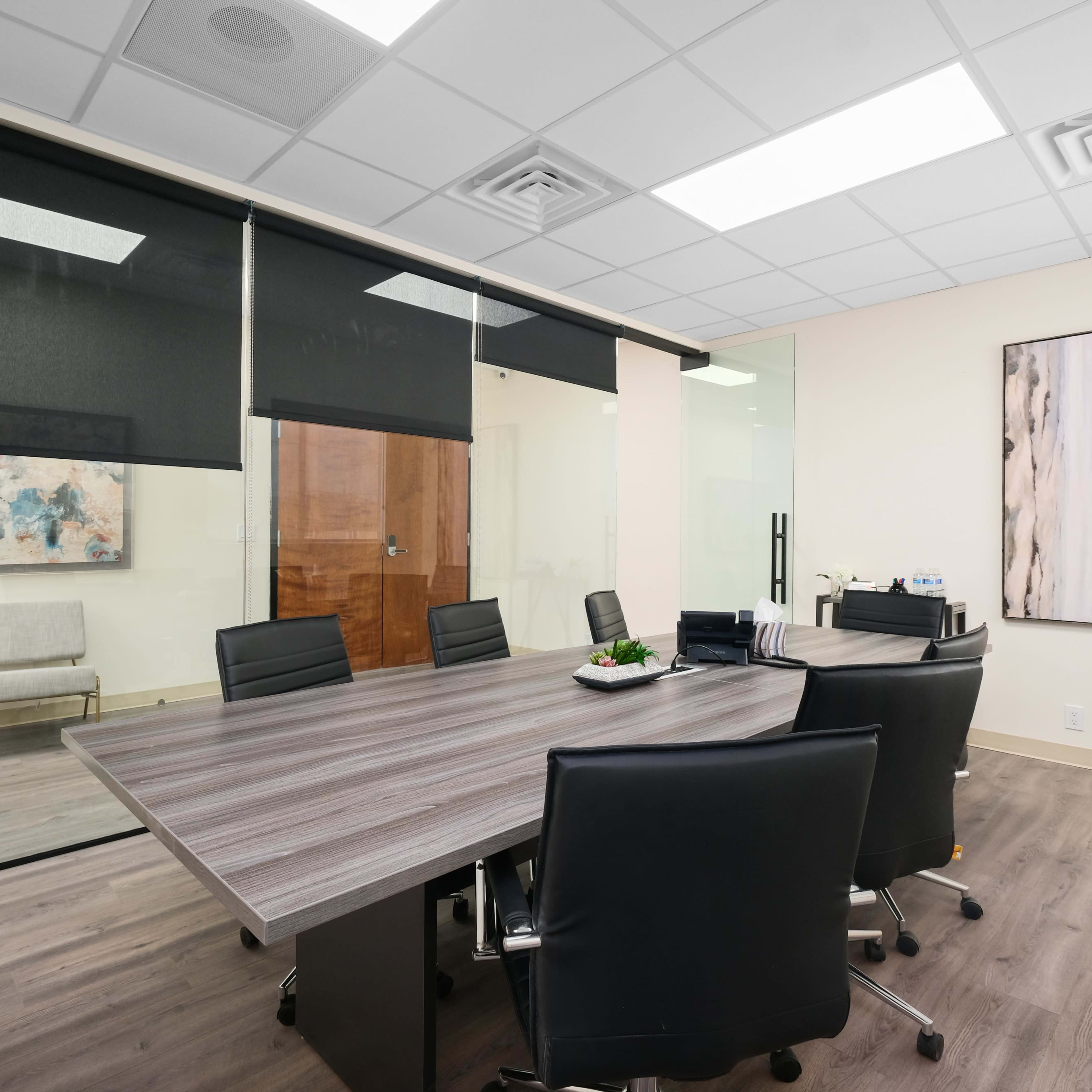 A modern office meeting room with a large table surrounded by black chairs, glass walls, and abstract artwork on the adjacent walls.