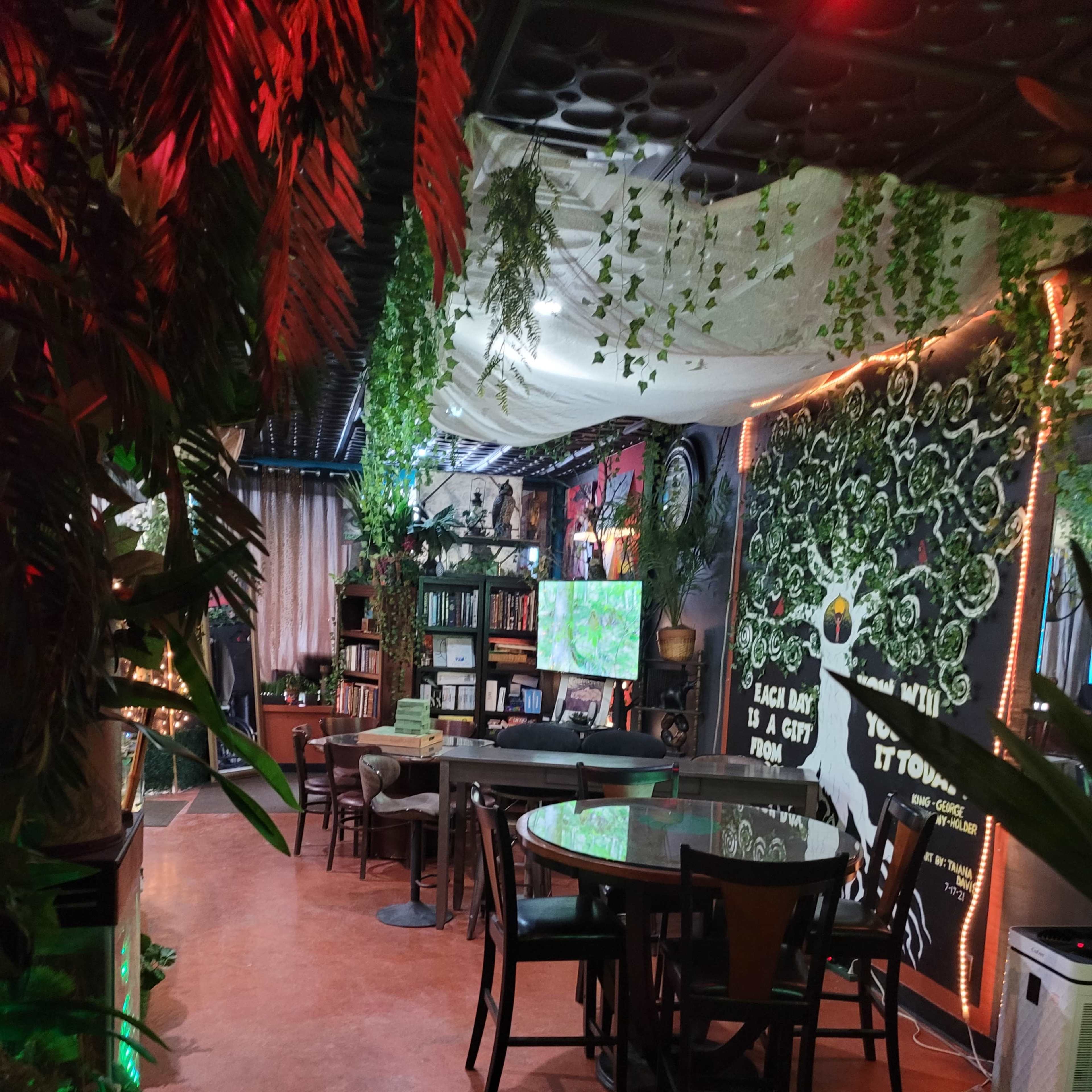 A brightly lit café with greenery, bookshelves, and a large wall mural.
