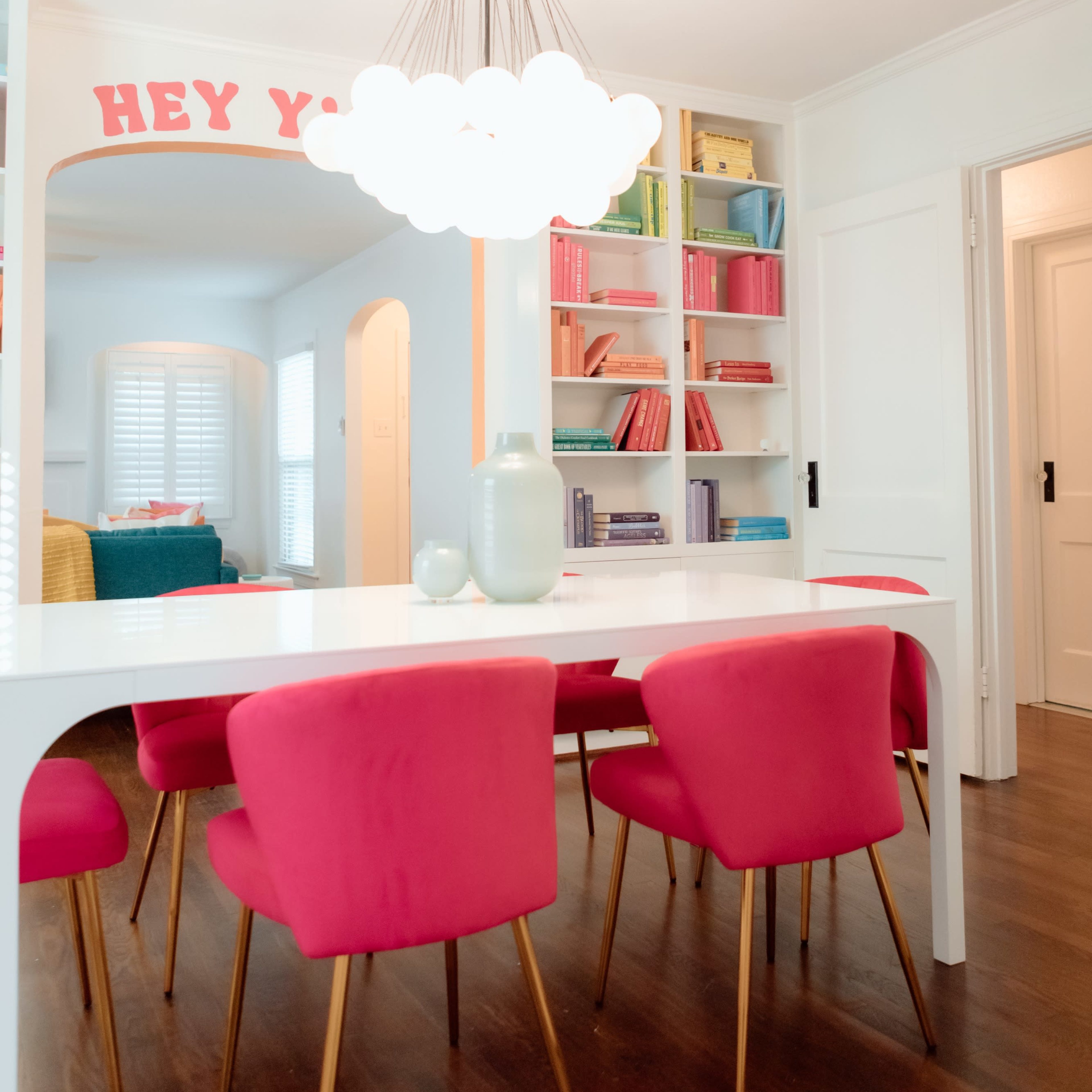 A brightly lit dining area features a white table surrounded by pink chairs and a colorful bookshelf in the background.