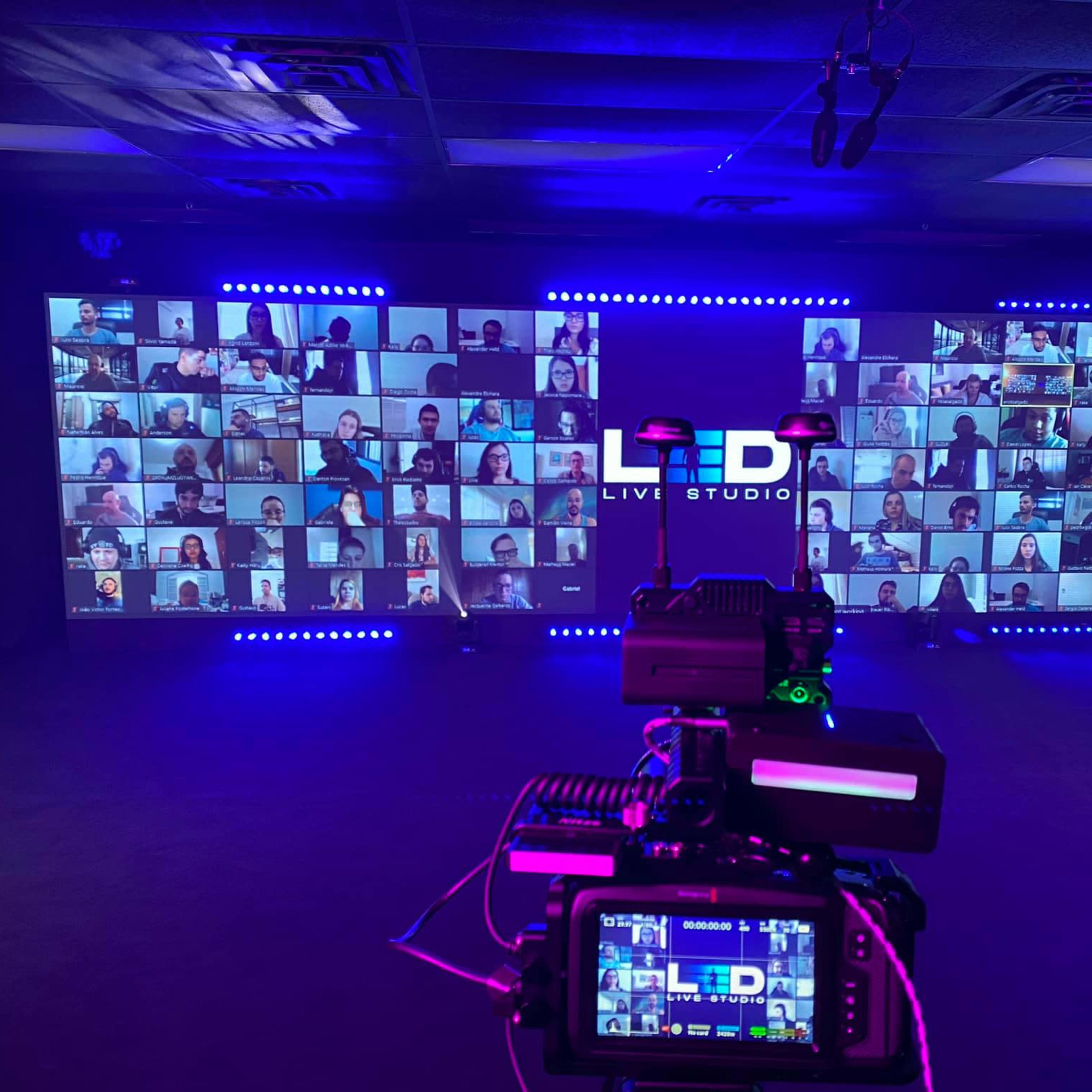 A camera captures a virtual event setup with numerous video conference participants displayed on large screens in a dimly lit studio.