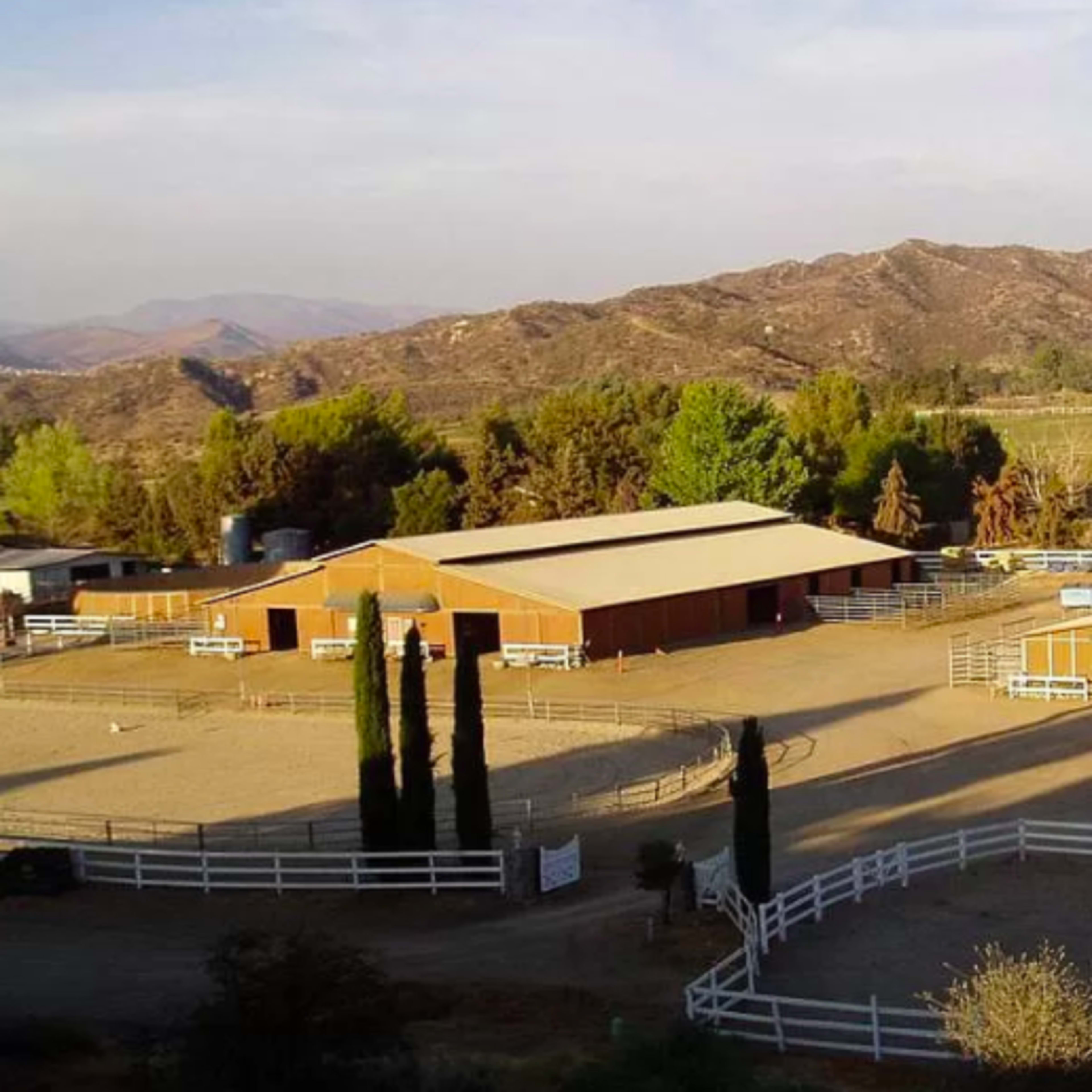 A sprawling equestrian facility features several wooden barns and riding areas, surrounded by mountains and trees.