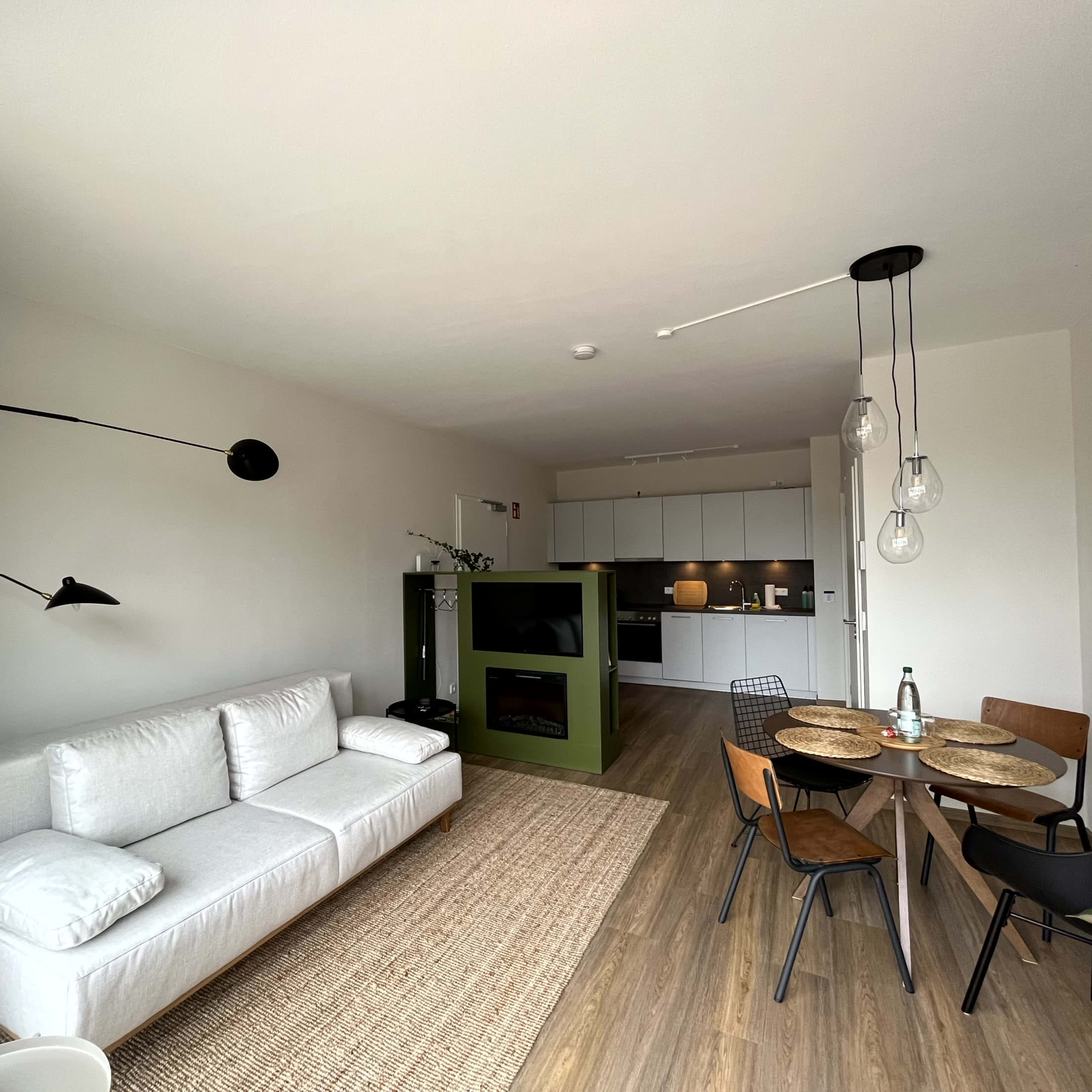 A modern living space featuring a white sofa, a dining area with four chairs, and a kitchen with minimalist cabinetry.