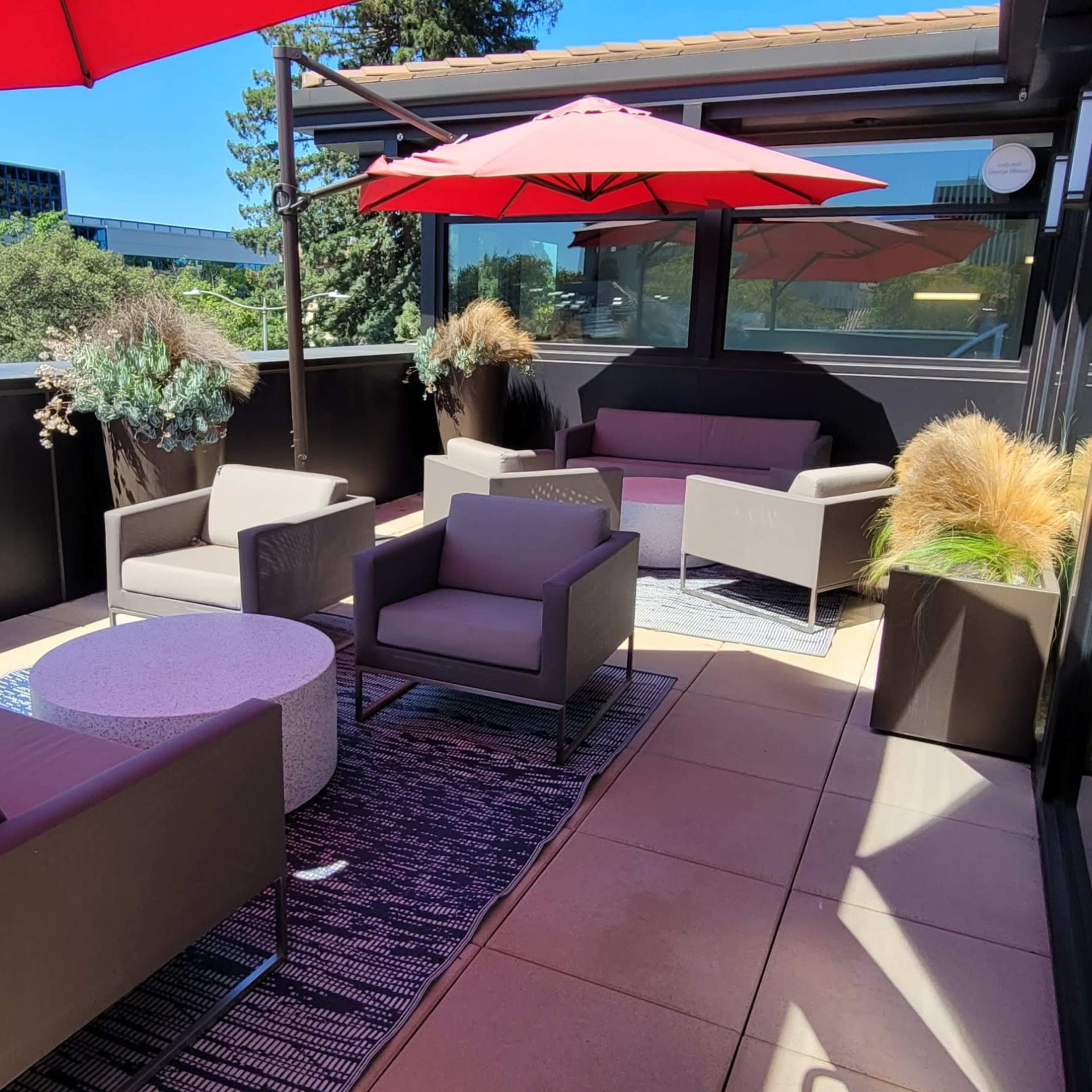 A rooftop terrace with several modern seating arrangements, a circular table, and red umbrellas providing shade.