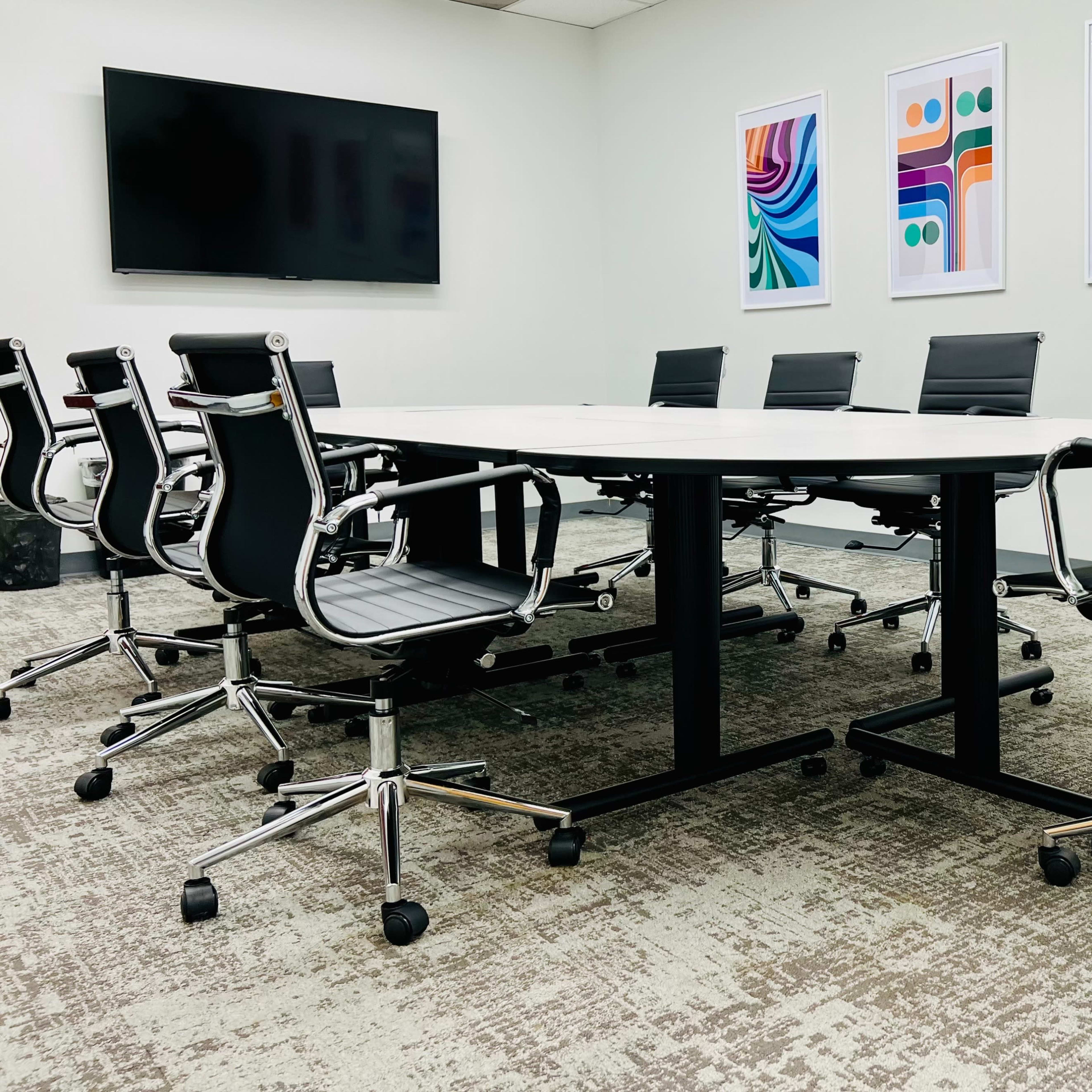 A conference room features a large round table surrounded by several black rolling chairs, with colorful abstract art displayed on the walls.