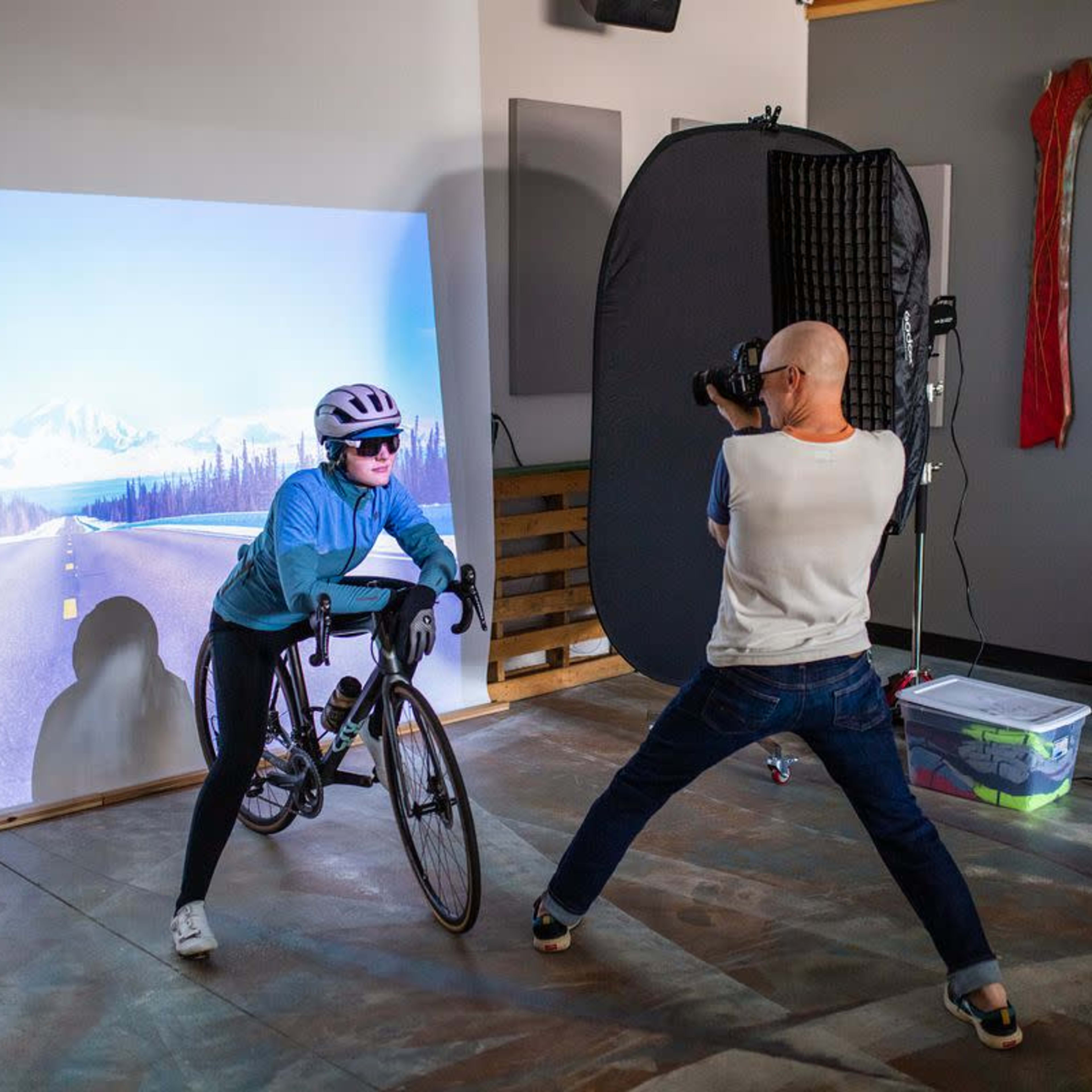 A cyclist poses on a stationary bike in a studio setting while a photographer captures the shot against a backdrop of a scenic road.