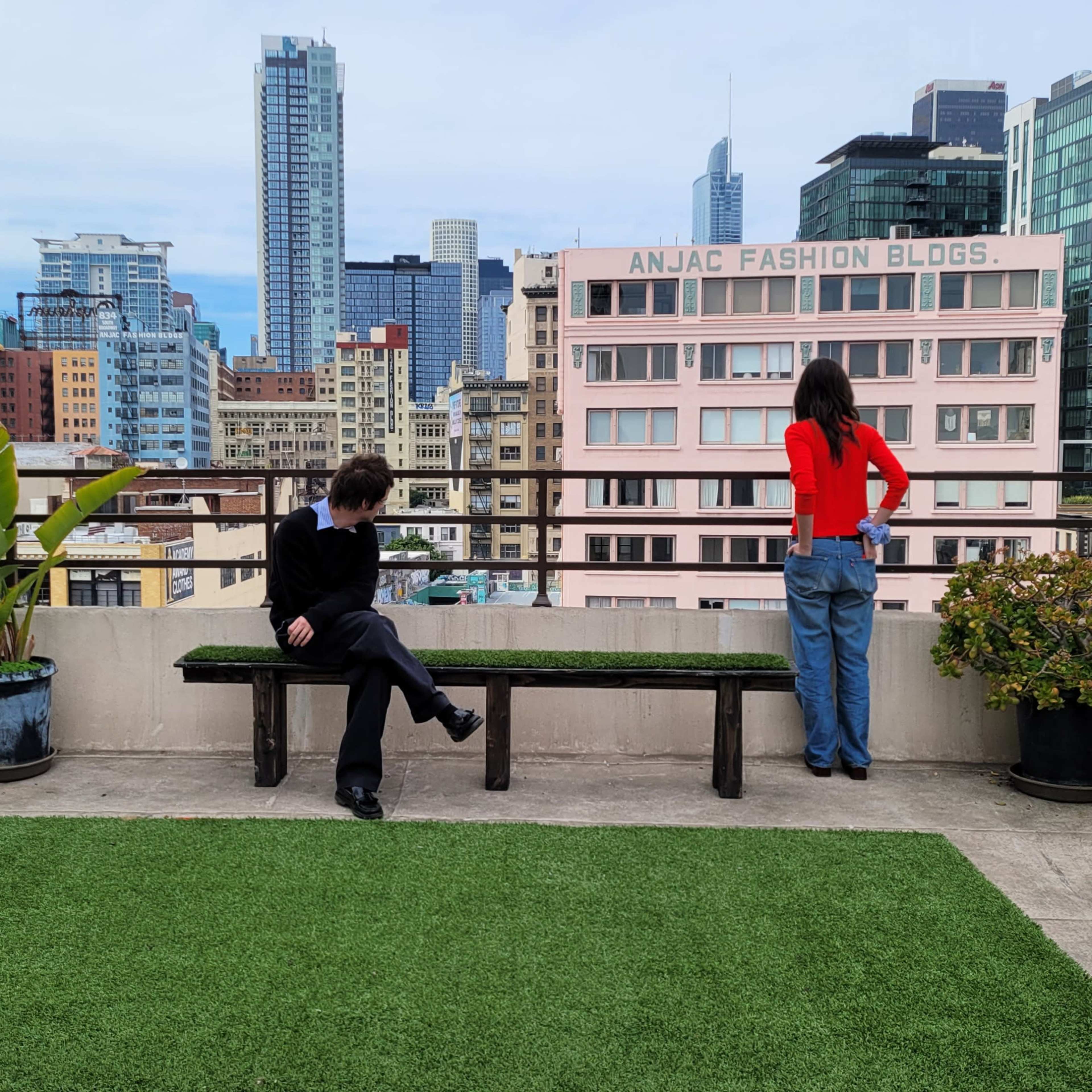 Two individuals are standing and sitting on a rooftop terrace, with a city skyline featuring tall buildings in the background.