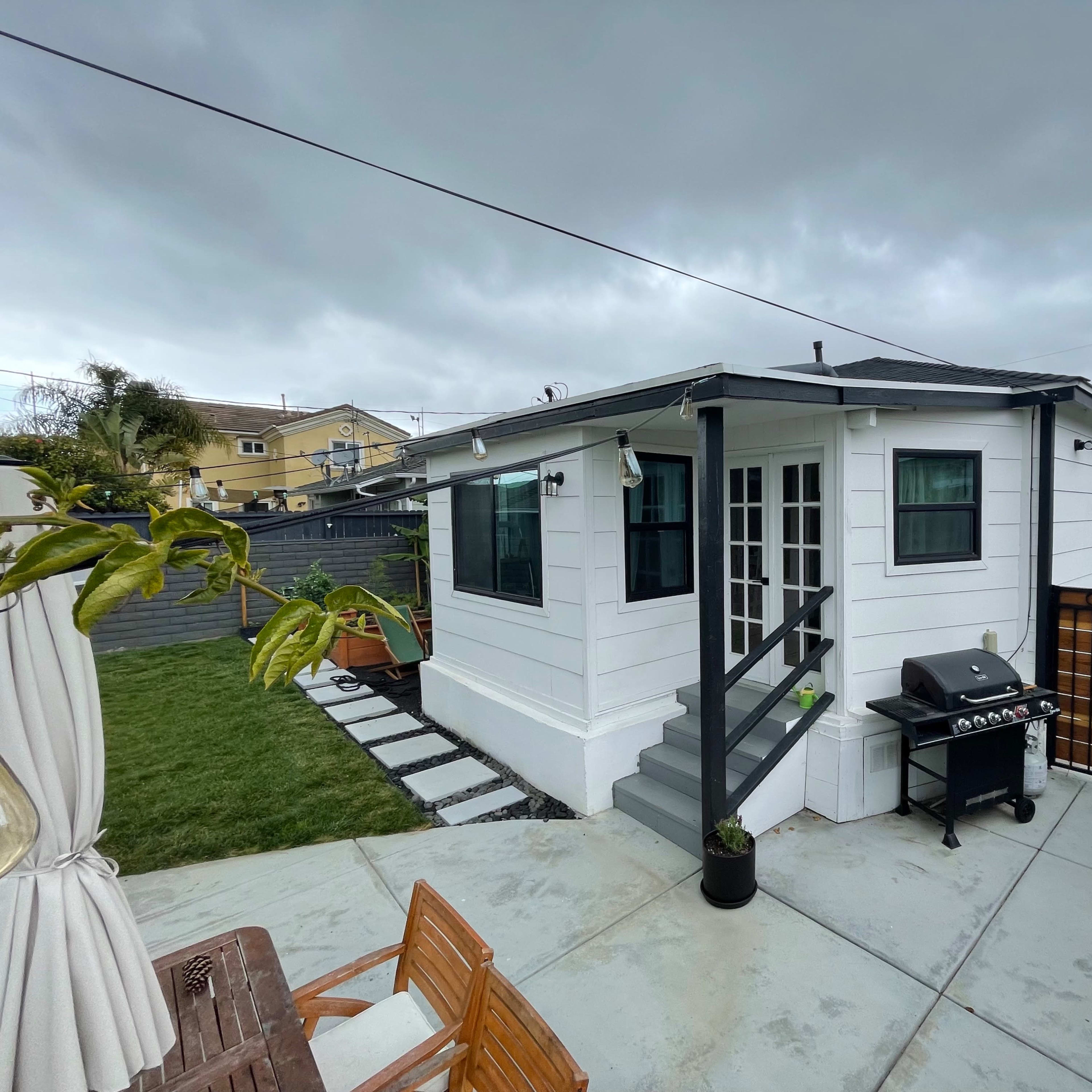 A small white house with a gray roof, a barbecue grill on a patio, and a grassy area surrounded by a wooden fence under a cloudy sky.