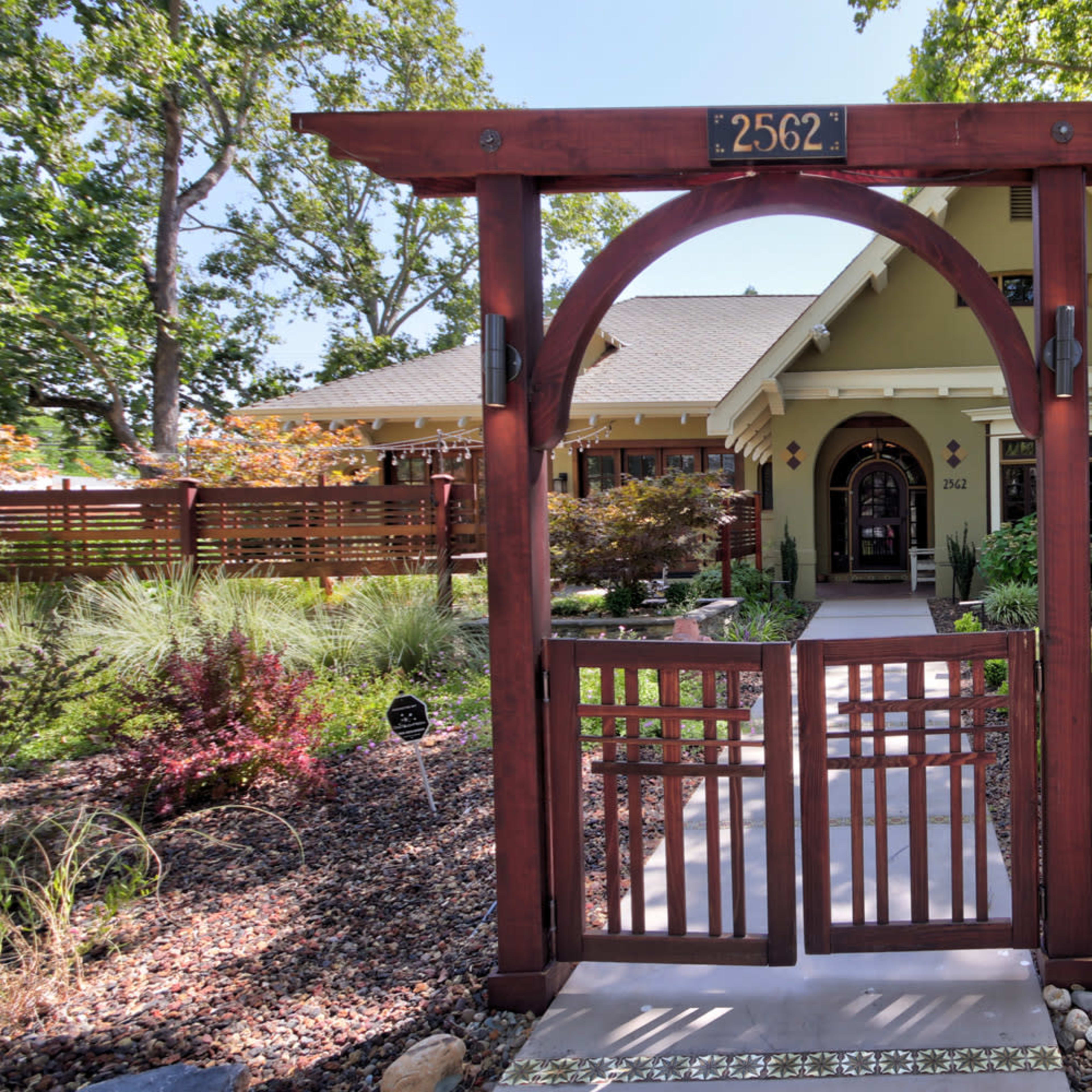 A wooden archway with a gate leads to a house surrounded by greenery and decorative landscaping.