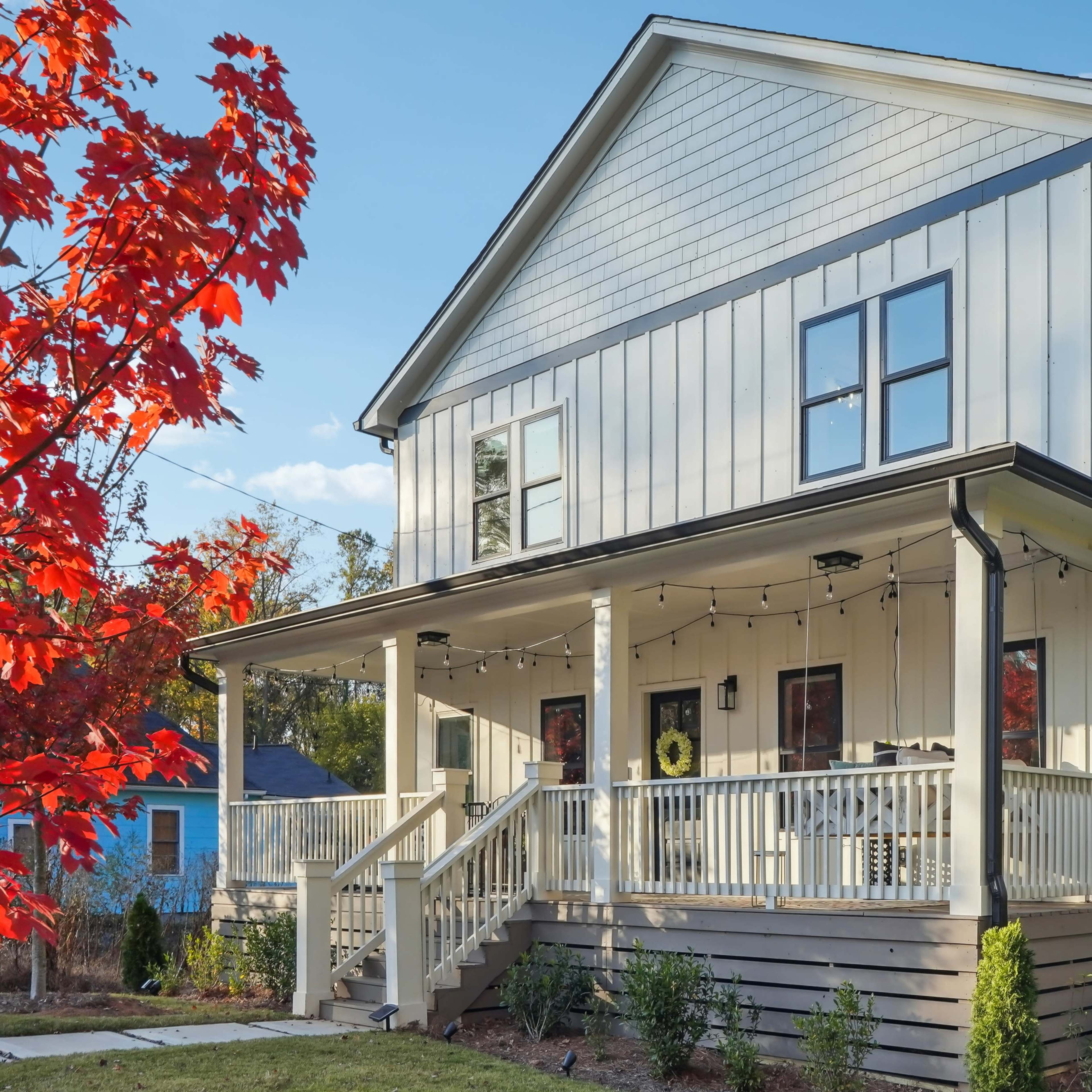A two-story, light-colored house features a spacious front porch with string lights and landscaped greenery.