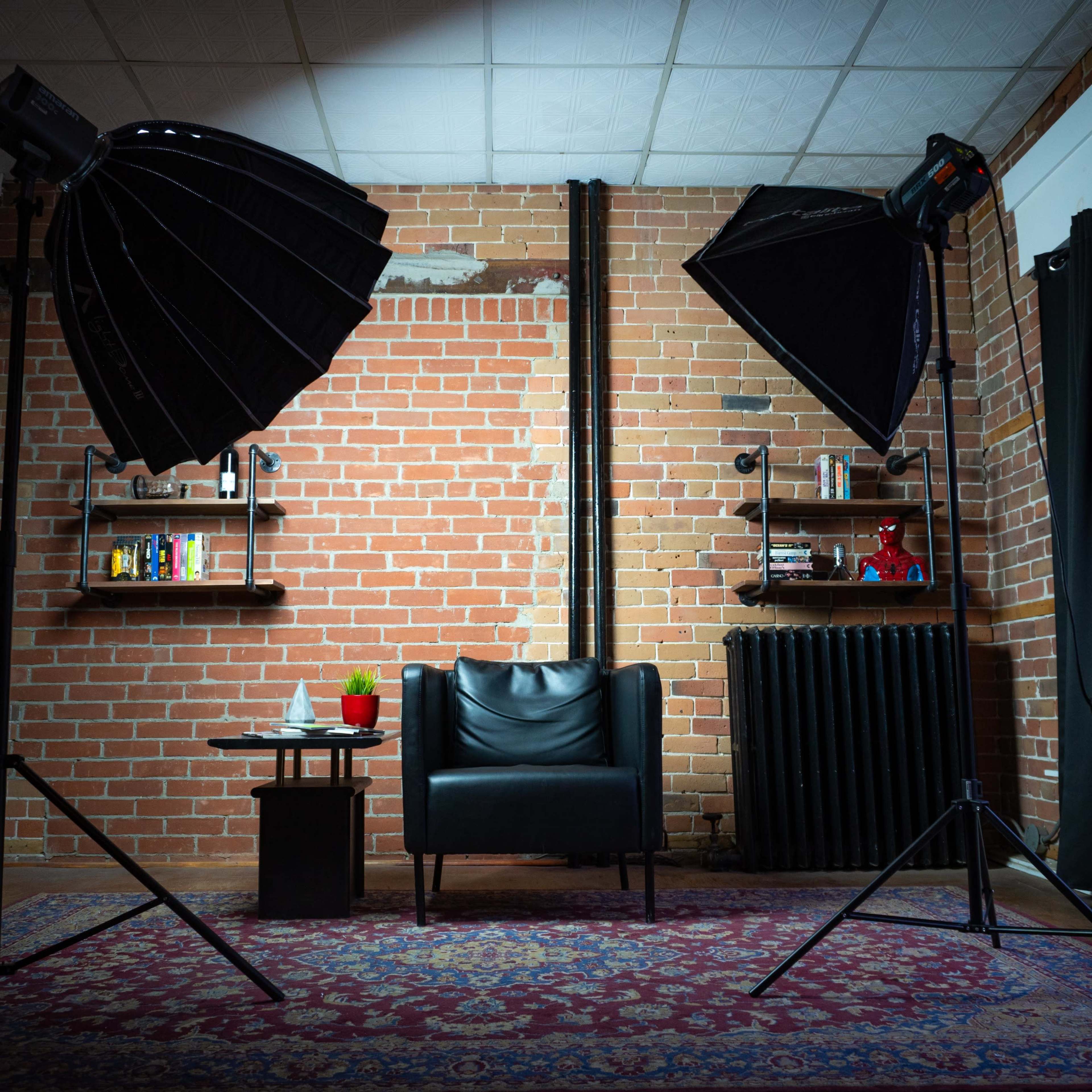 A well-lit studio setup featuring a black chair, a small table with drinks, and two softbox lights positioned on either side against a brick wall.
