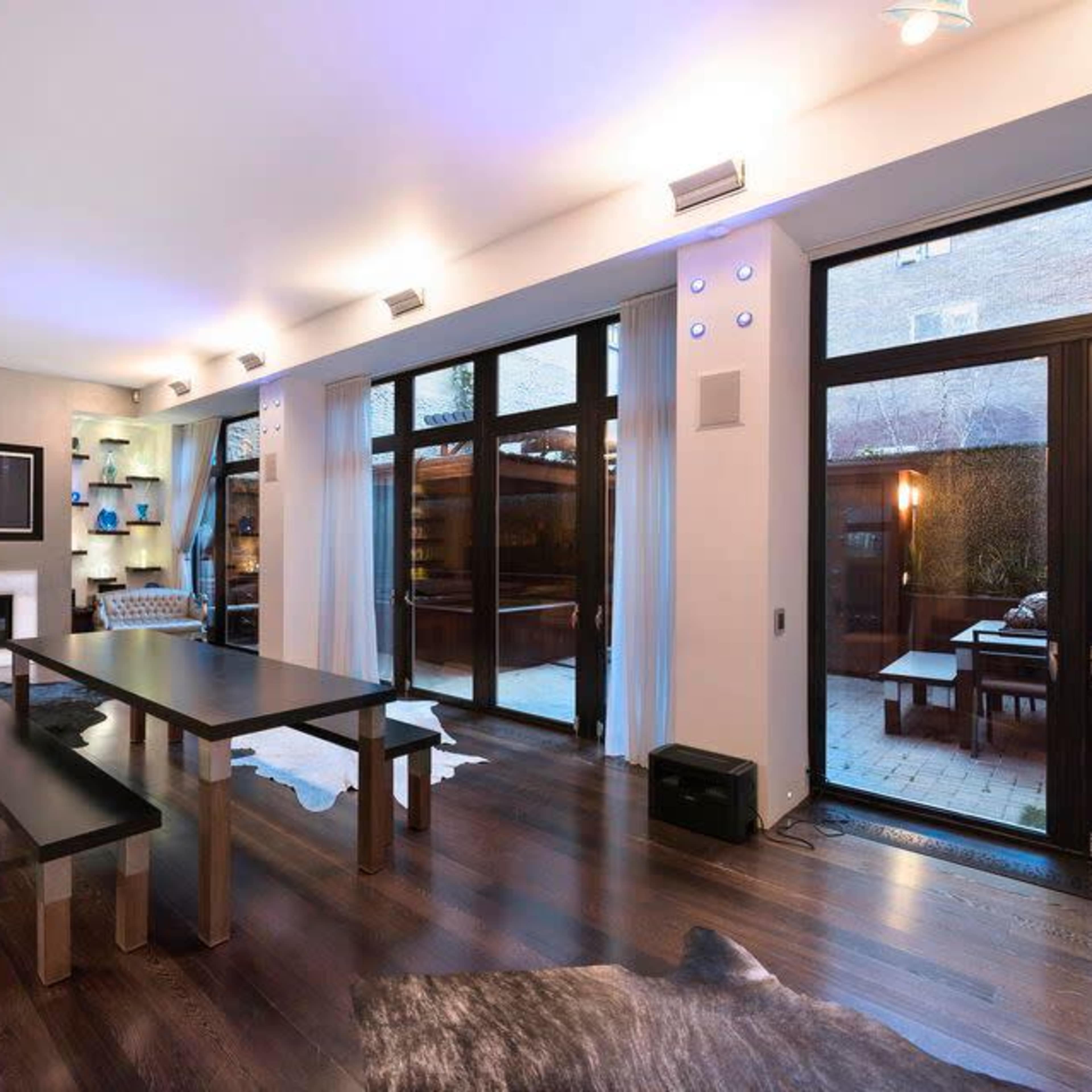 A modern living room with large windows, a flat-screen TV mounted on the wall, and a wooden table set on a dark hardwood floor.