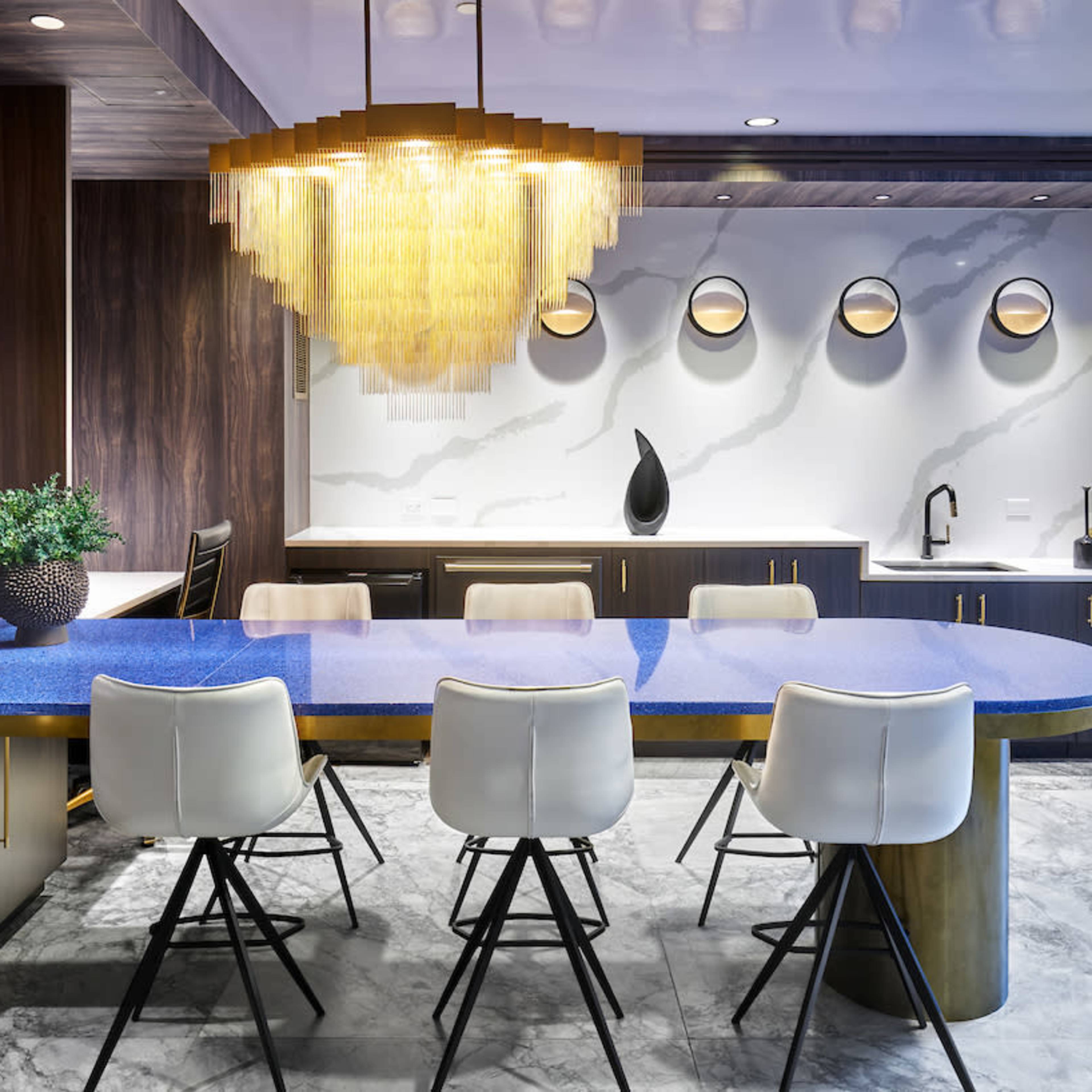 A modern kitchen features a blue countertop with a circular table surrounded by white chairs, against a backdrop of dark cabinetry and decorative wall elements.