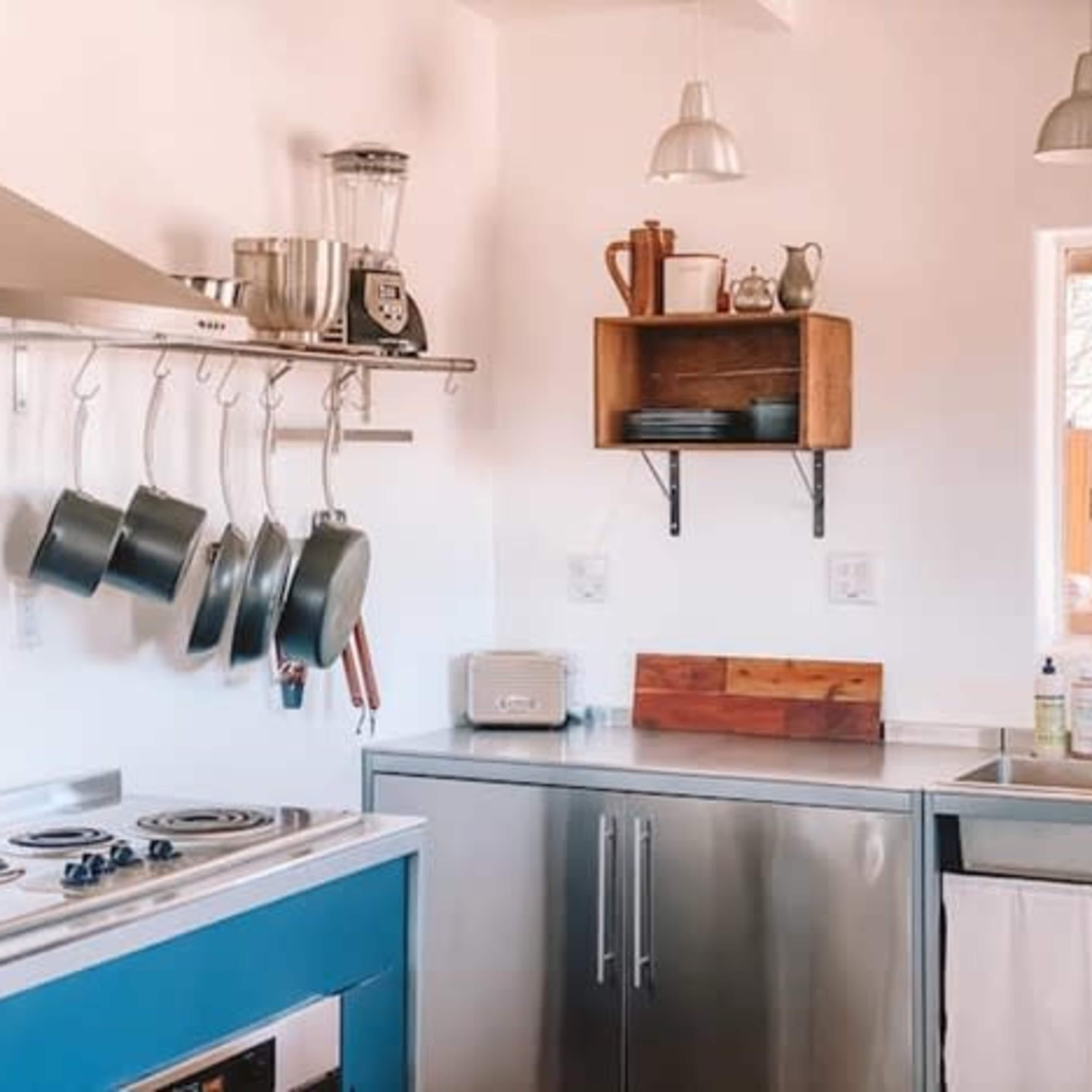 The kitchen features a stainless steel counter, a hanging rack with pots, and a window that provides natural light.