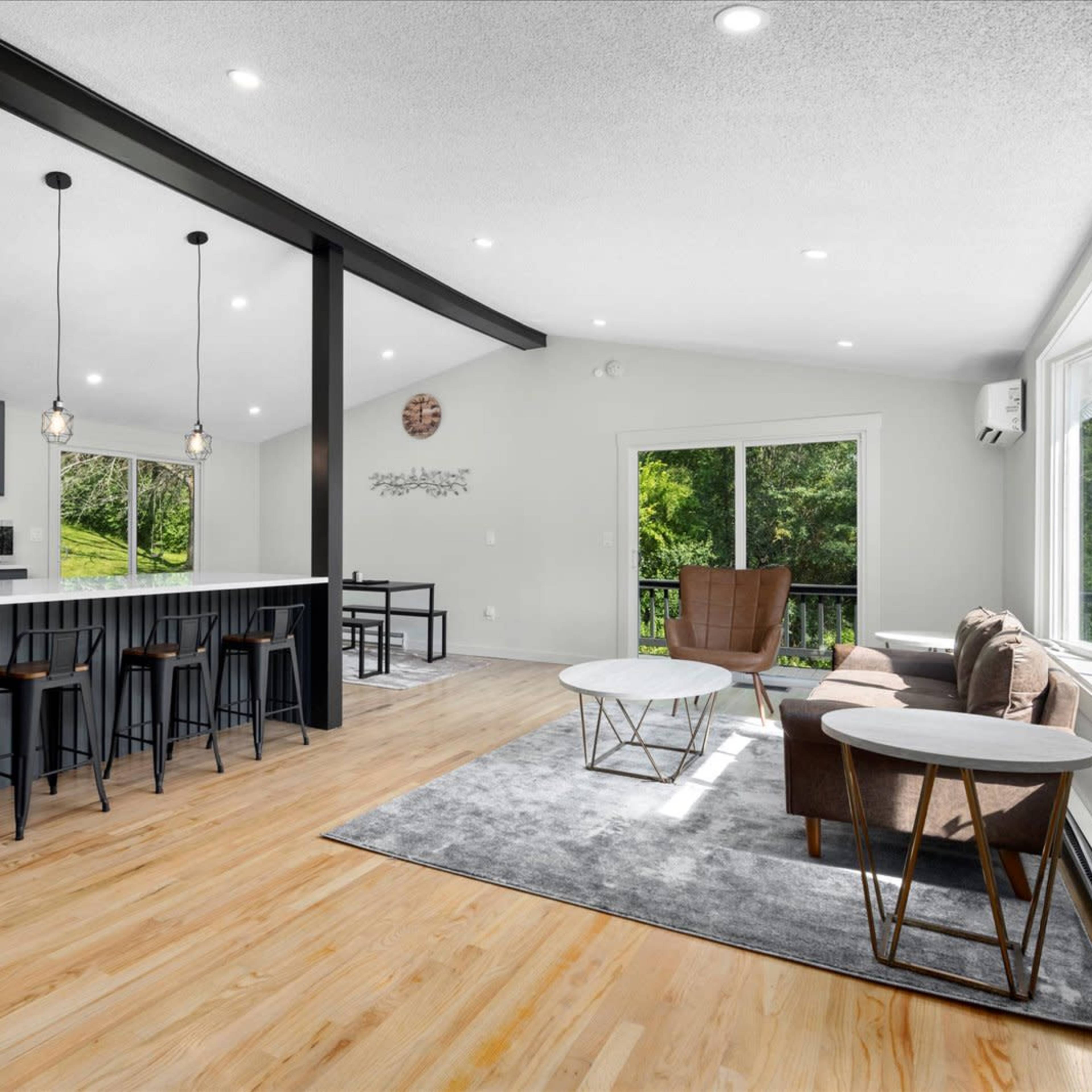 A spacious, modern living area with a kitchen island, bar stools, and large windows overlooking greenery.
