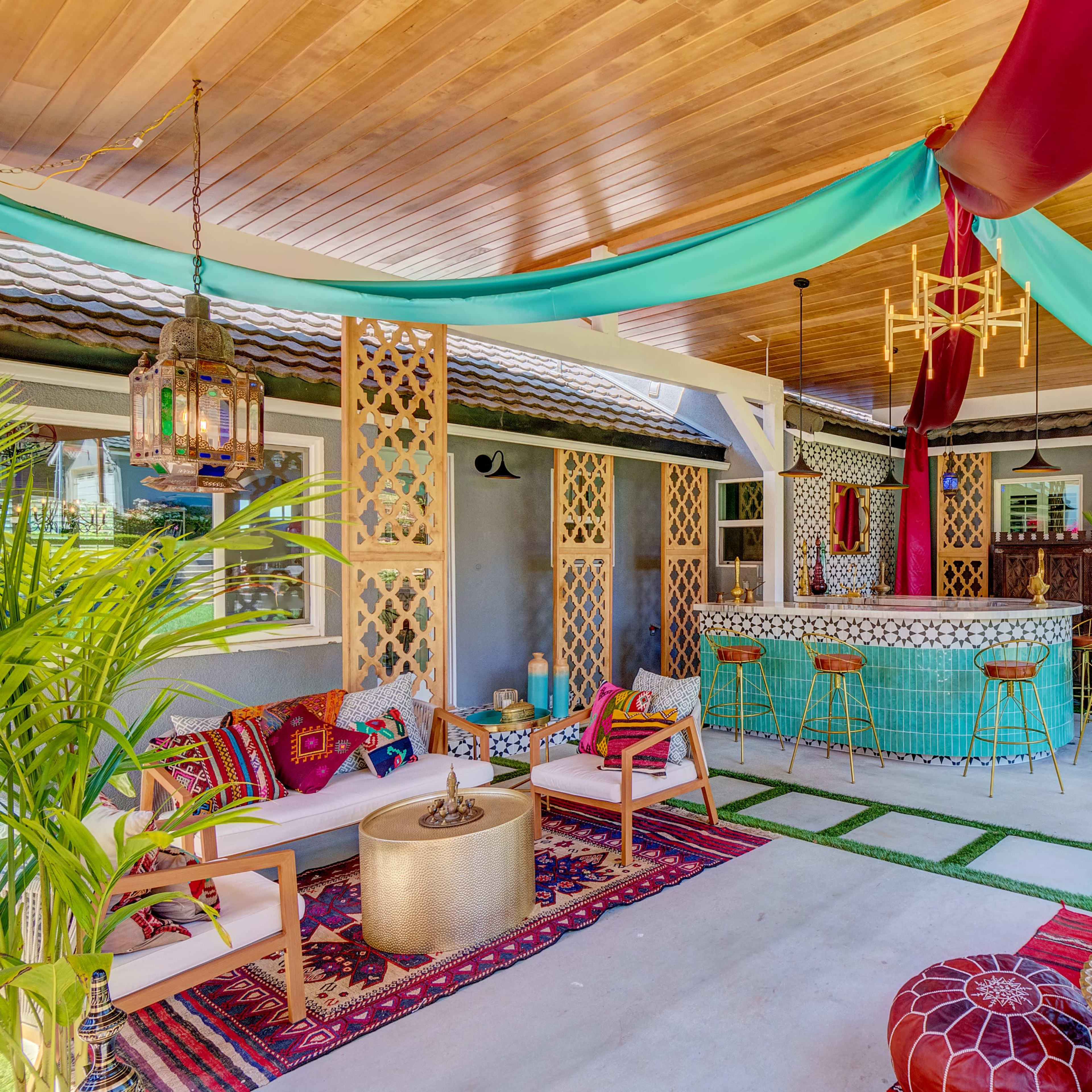 A colorful outdoor seating area features comfortable furniture, decorative fabrics, and a bar surrounded by plants.