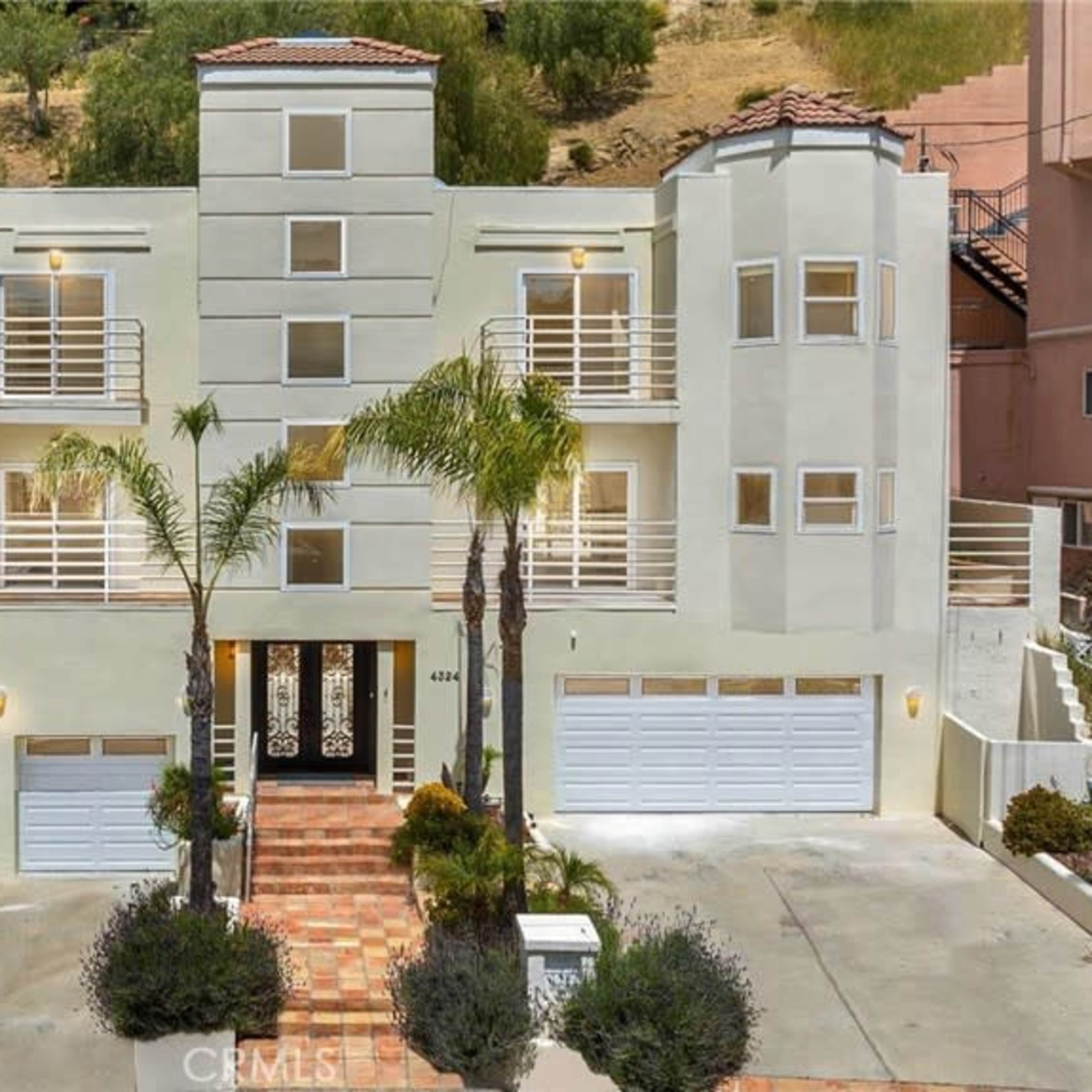 A three-story, modern house with a landscaped front yard, three garage doors, and a distinct architectural style.