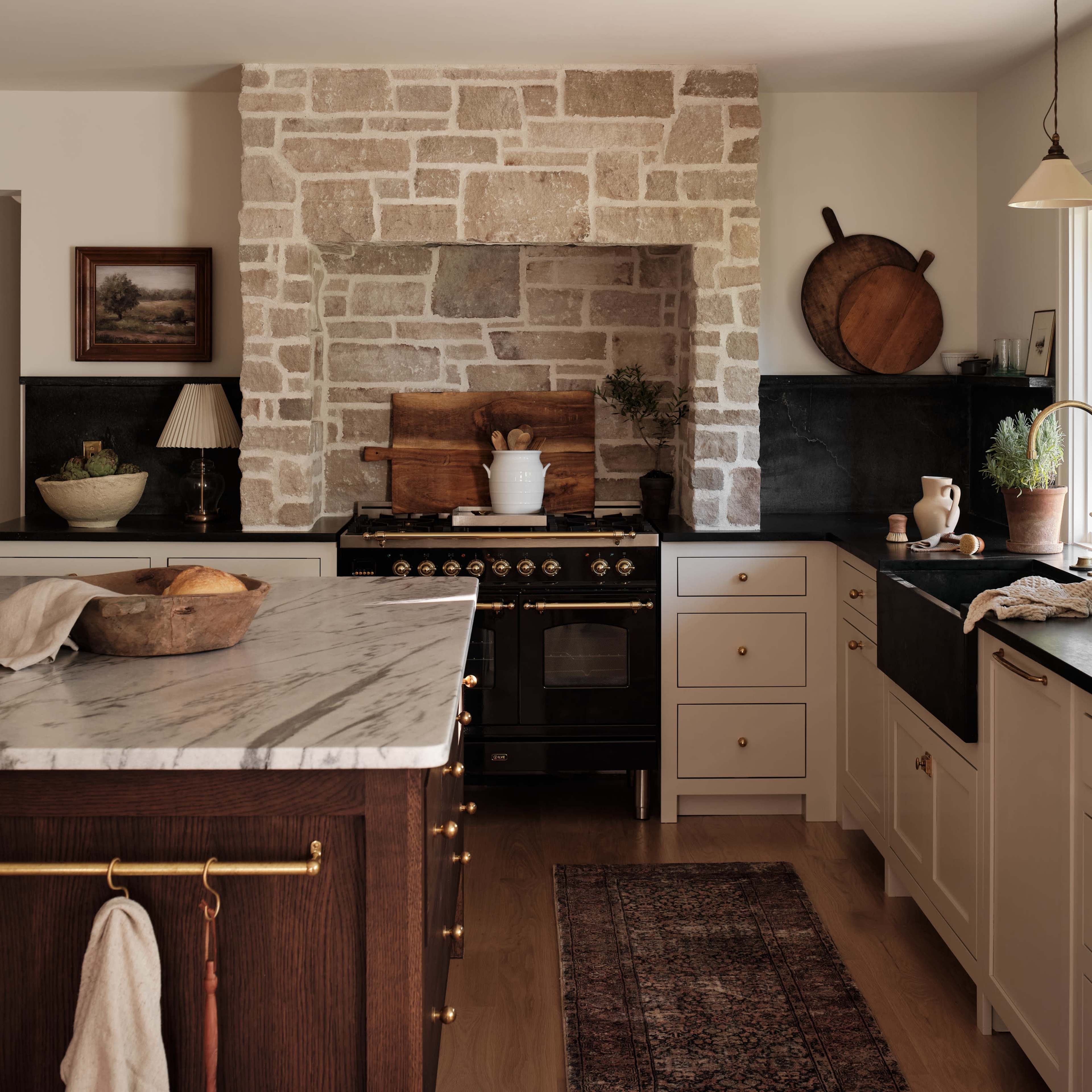 A modern kitchen features a large marble island, stone wall, and dark cabinetry with brass accents.