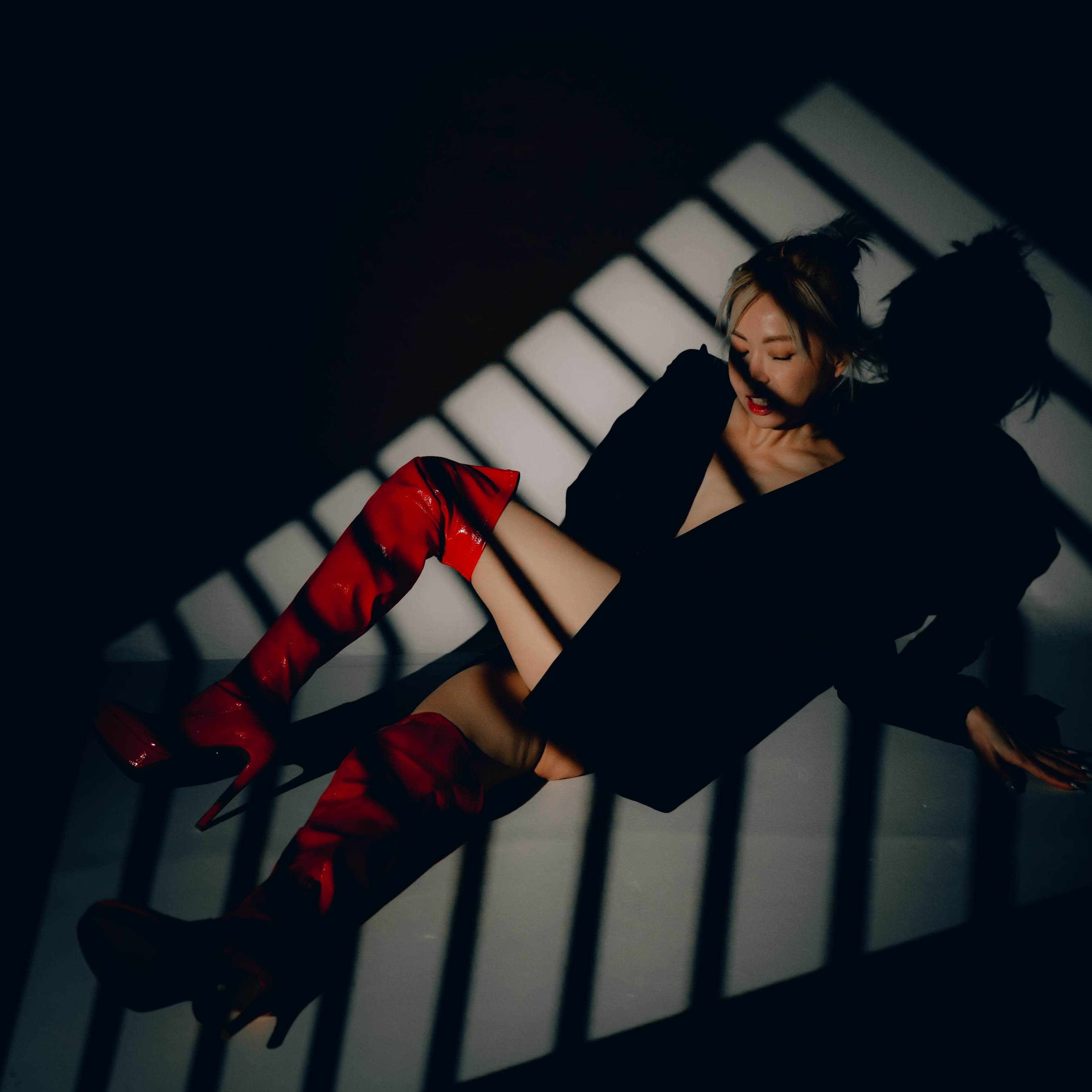 A person wearing a black outfit and red thigh-high boots lies on the ground, casting a shadow against diagonal light stripes.