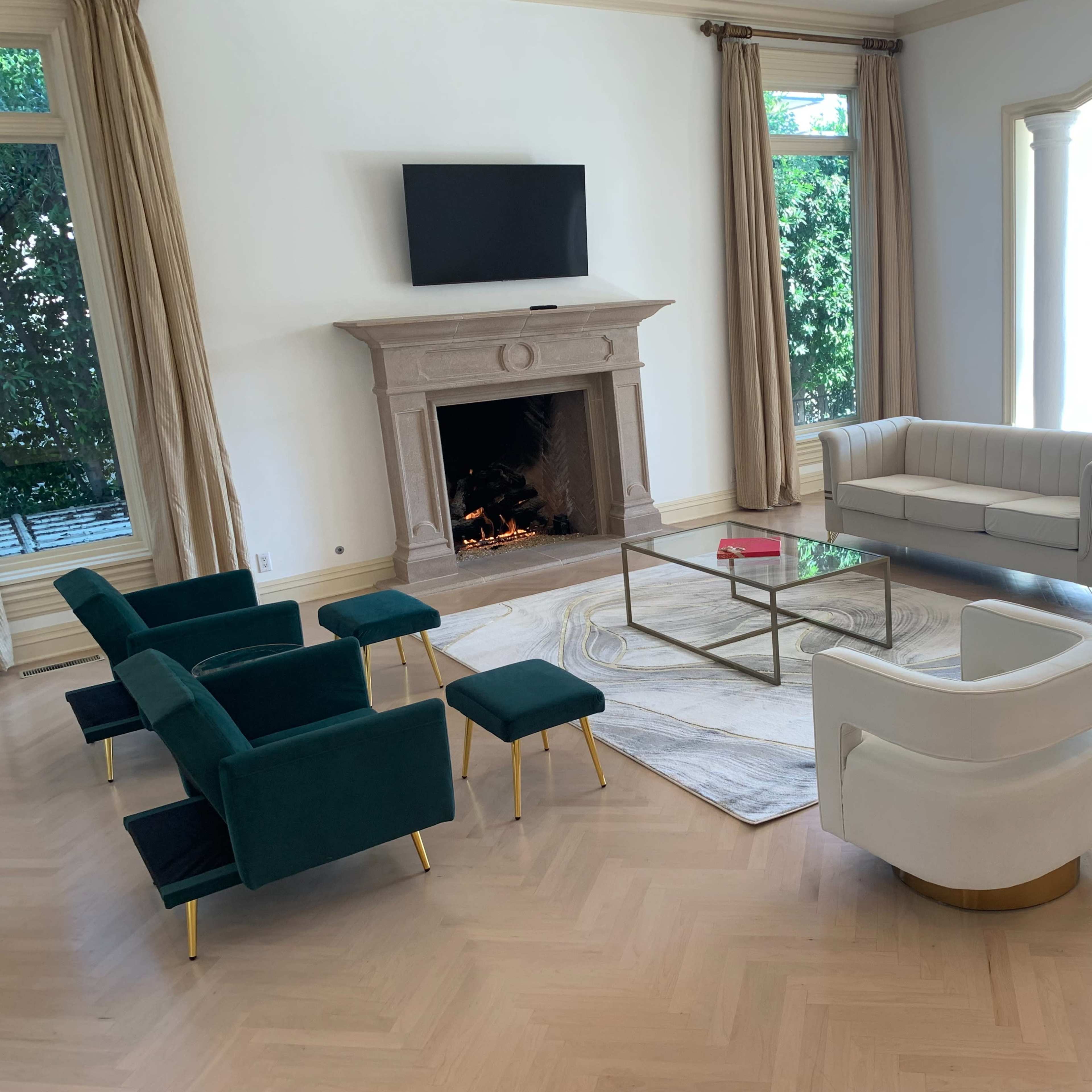 The living room features a modern setup with a white sofa, two curved chairs, green accent chairs, a glass coffee table, and a stone fireplace, all arranged around a light-colored area rug.