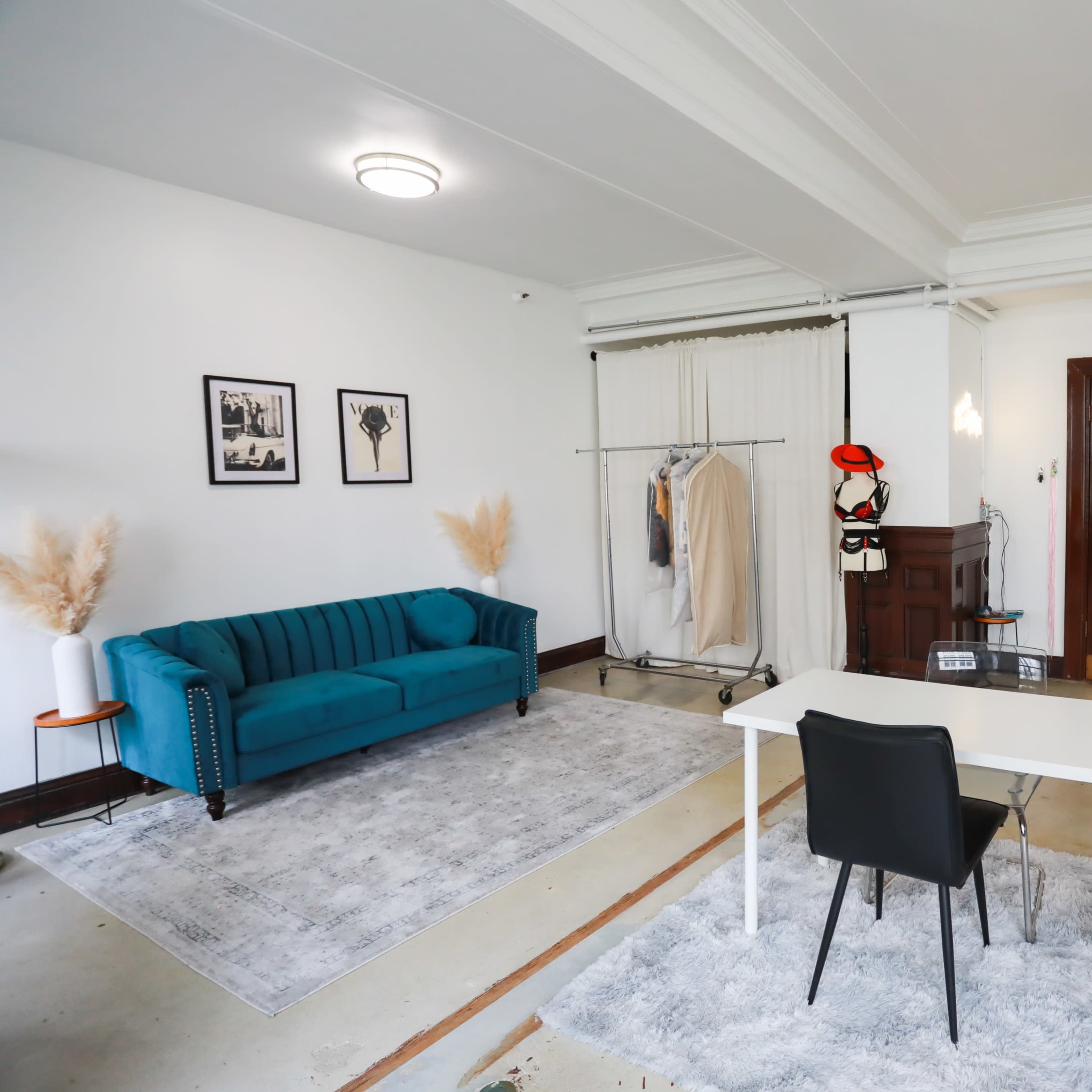 A well-lit room featuring a teal velvet sofa, a clothing rack with garments, a white table with two black chairs, and decorative elements like pampas grass.
