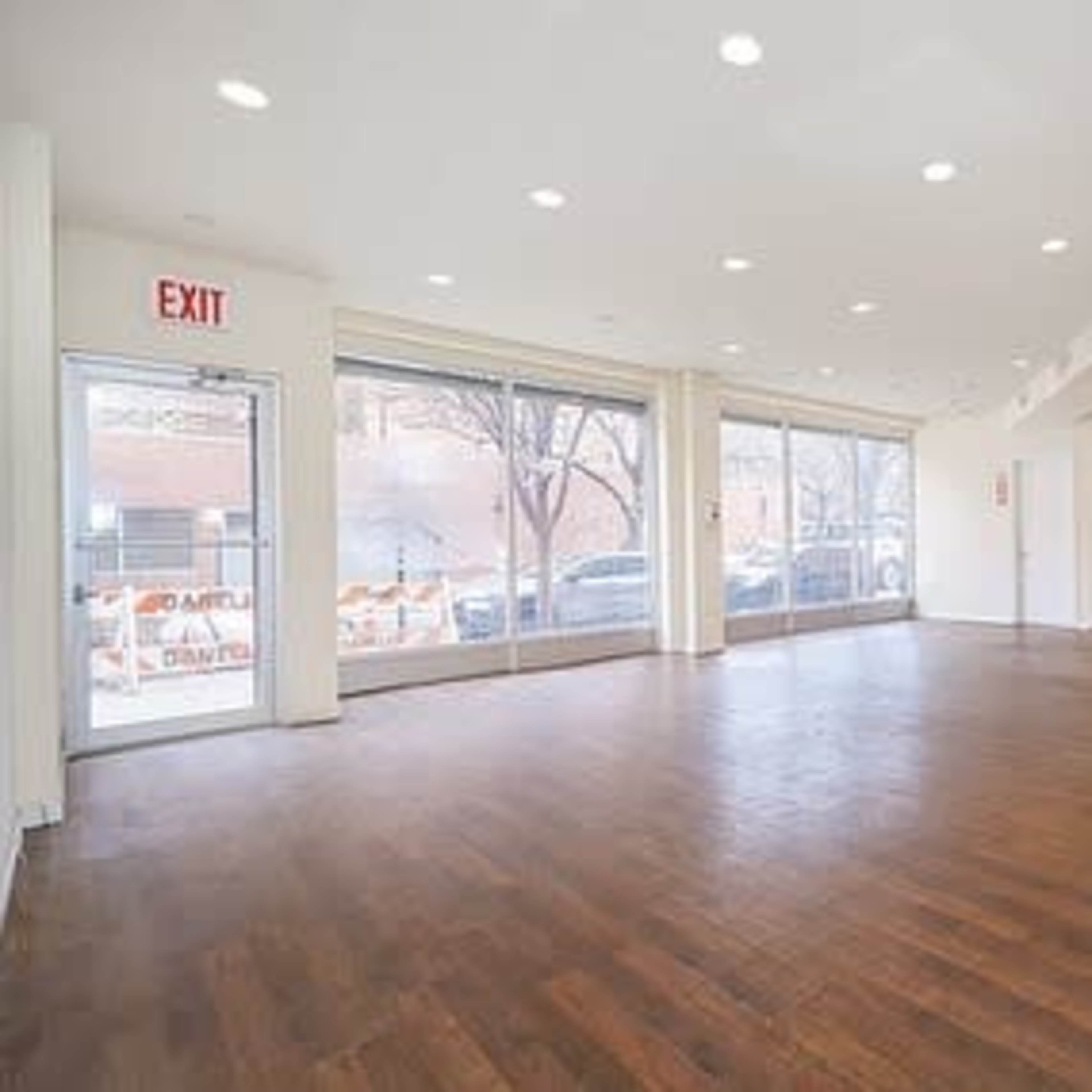 An empty retail space with hardwood flooring, large windows, and an exit sign near the entrance.