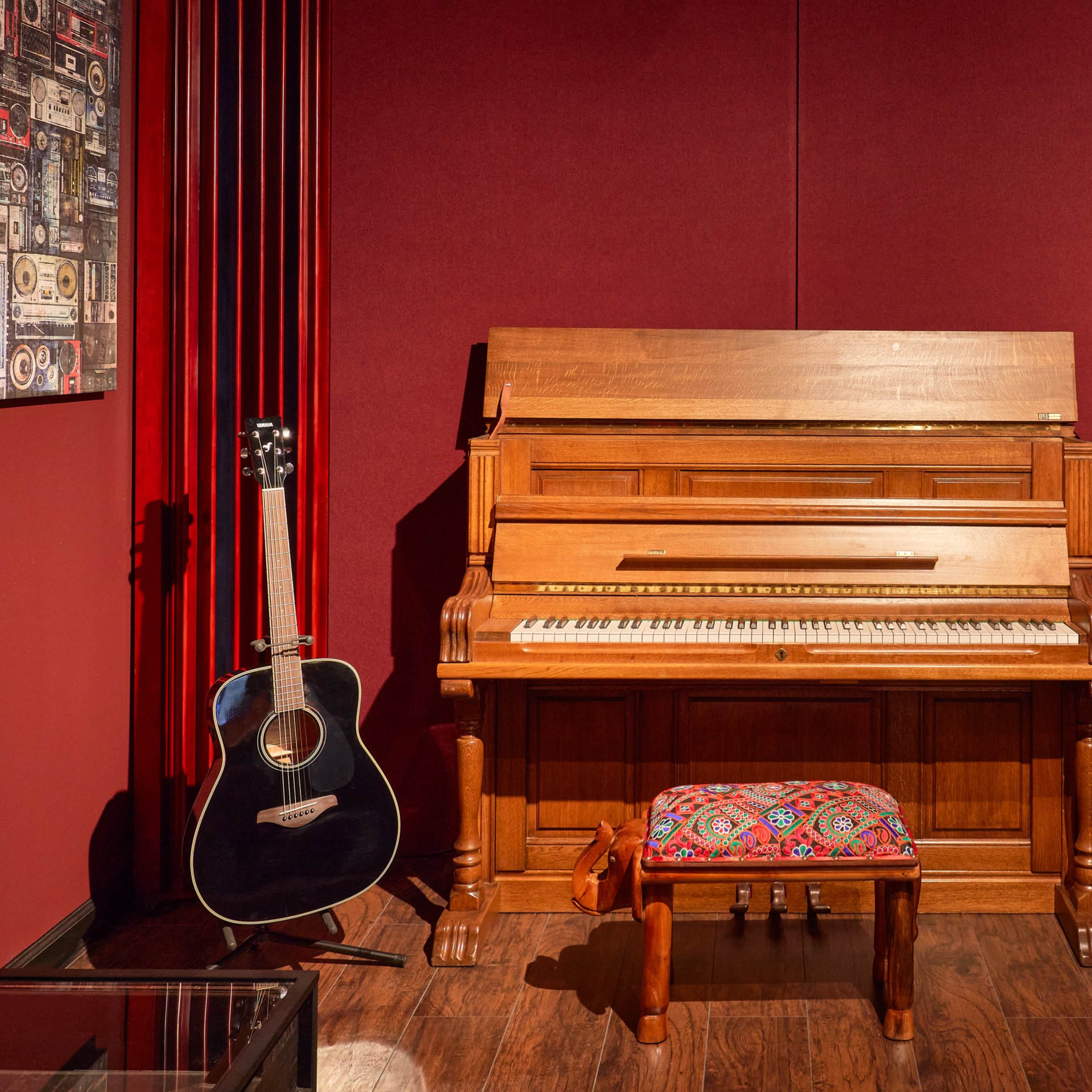 A vintage wooden piano sits against a red wall, accompanied by an acoustic guitar and a laptop on a small table.