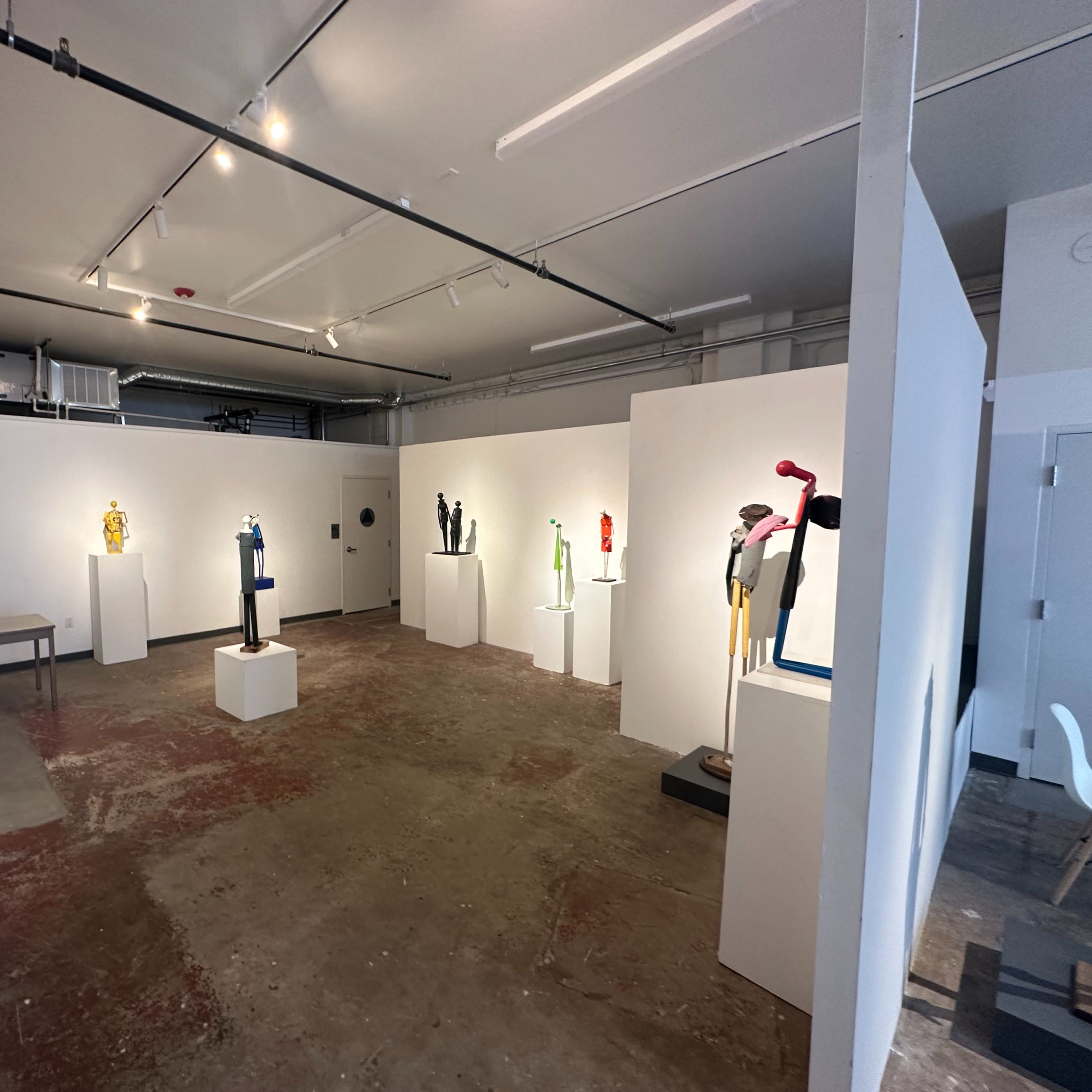 An art gallery featuring white pedestals displaying various sculptures against a backdrop of gray walls and a concrete floor.