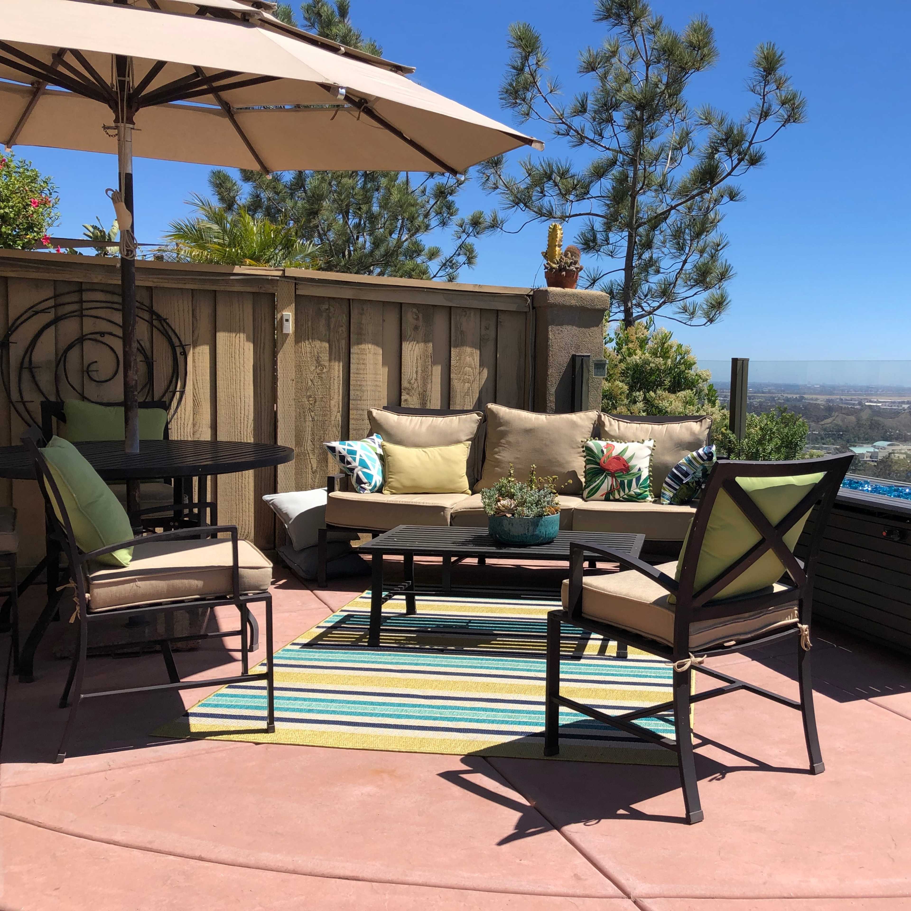 A patio area features a table, chairs, and a sectional sofa, surrounded by plants and with a view overlooking the landscape.