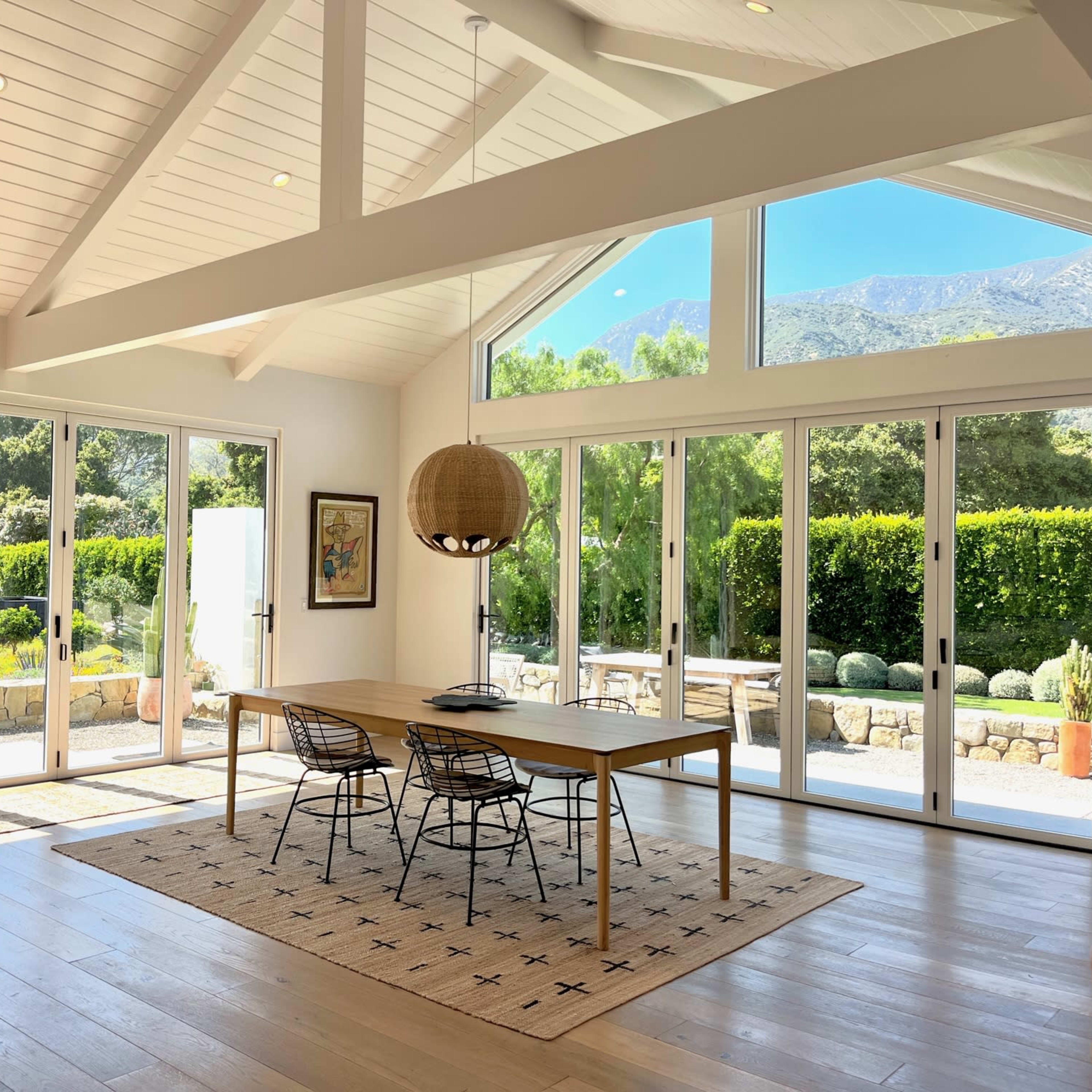A bright, airy dining area features a wooden table surrounded by black chairs, with large windows offering a view of a landscaped garden.