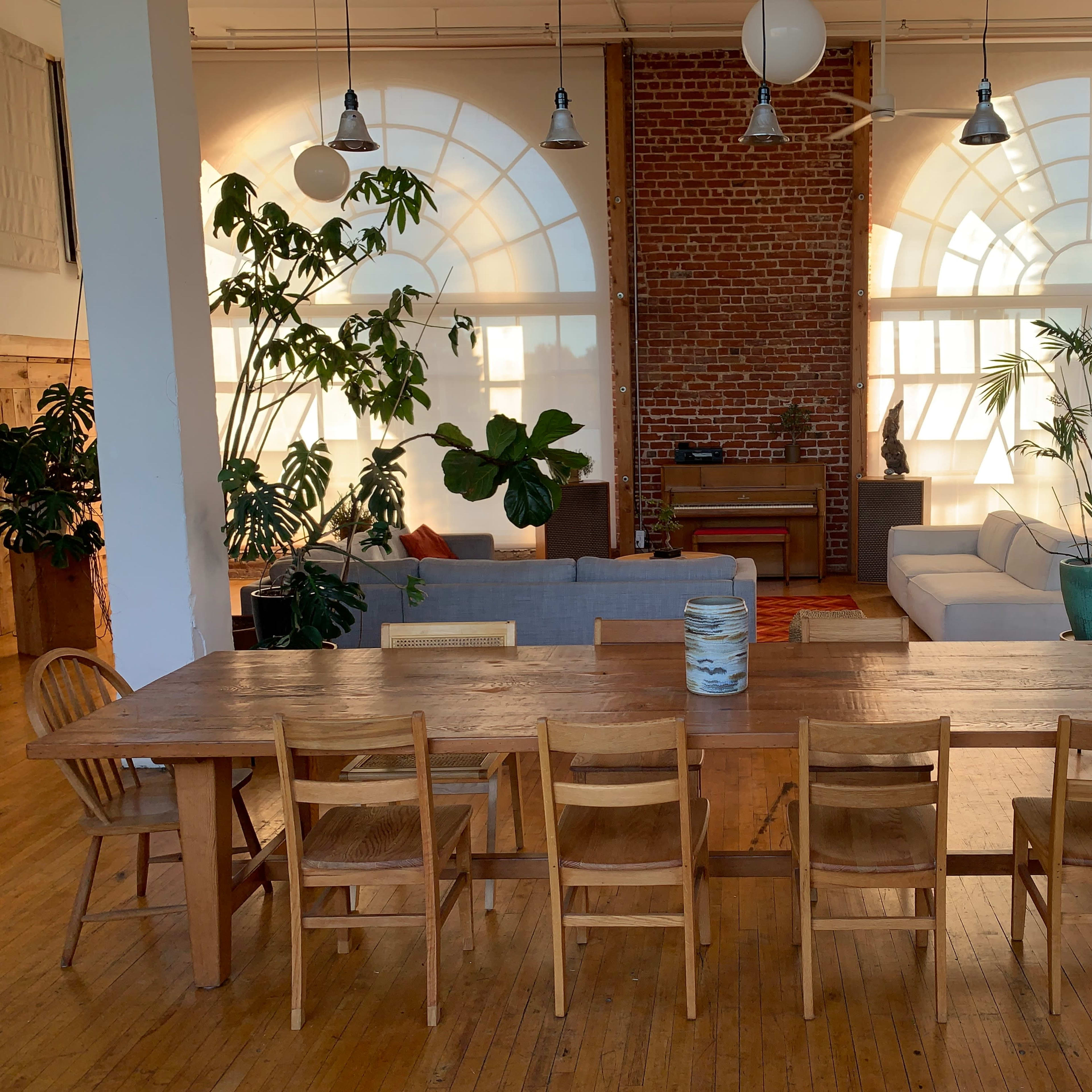 A long wooden dining table with multiple chairs is set in a spacious room featuring large windows and exposed brick walls, with various plants and a sofa in the background.
