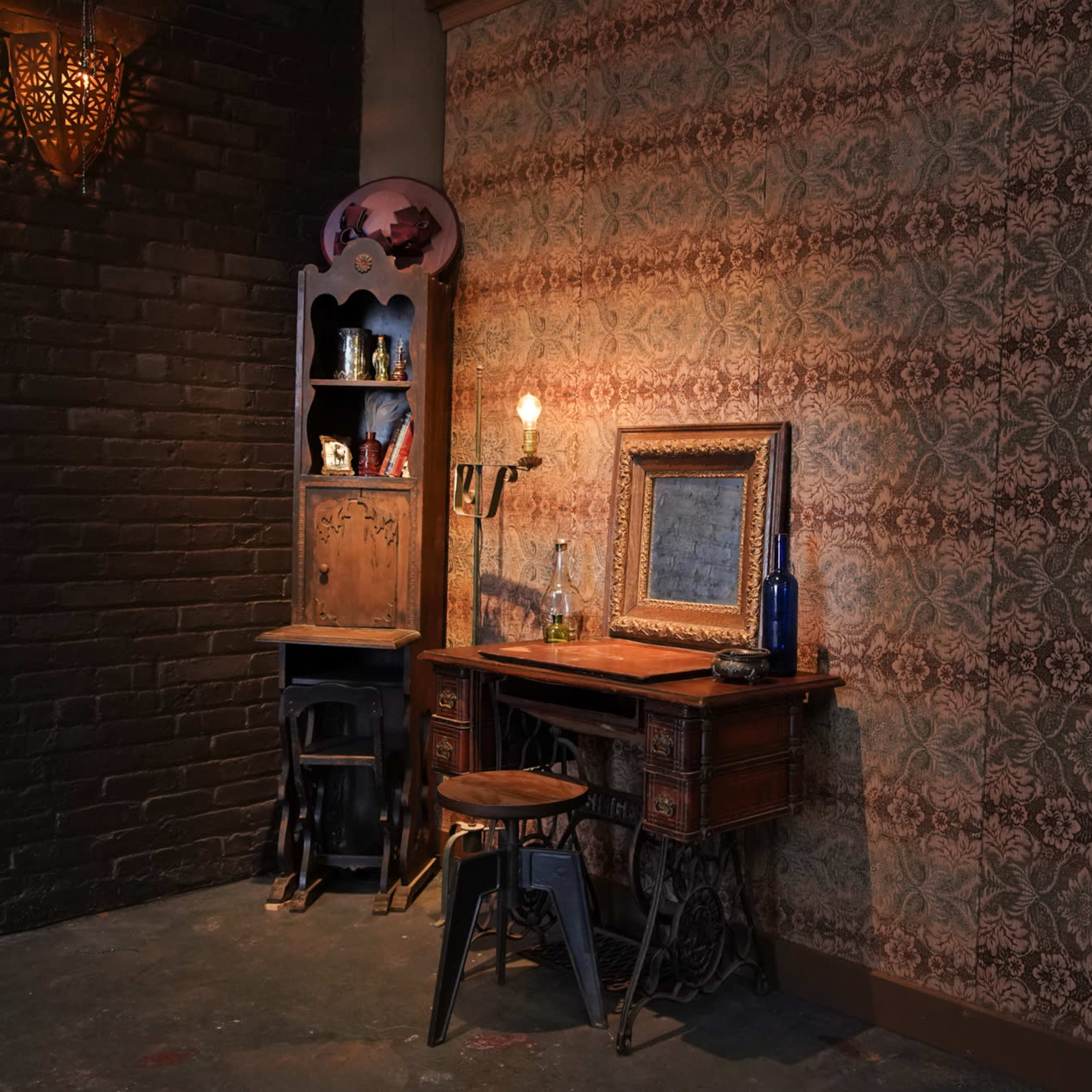 A vintage-style corner with a wooden desk, a framed mirror, a stool, and a small bookshelf against a patterned wallpaper backdrop.