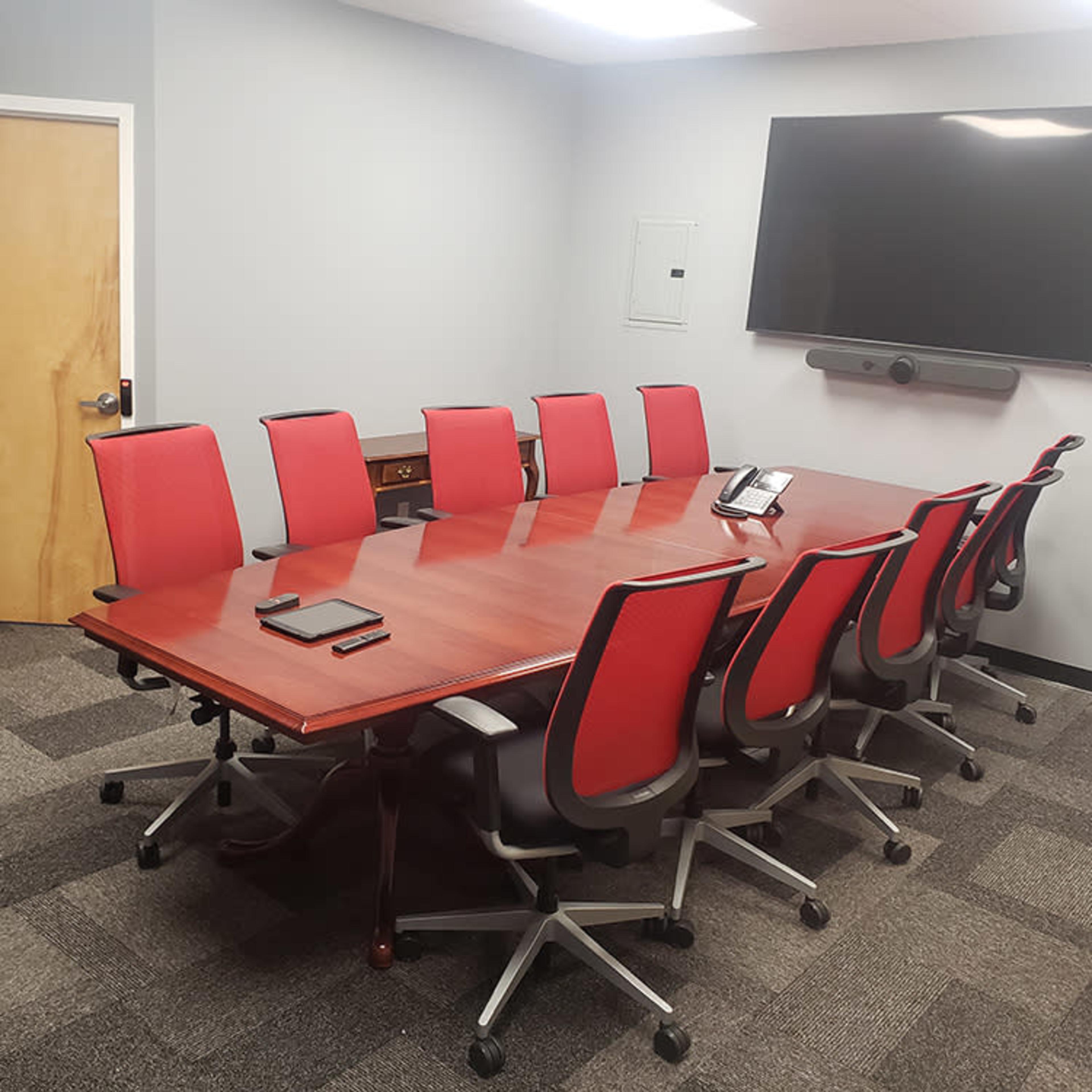 A conference room features a long wooden table surrounded by red office chairs, with a large screen mounted on the wall.