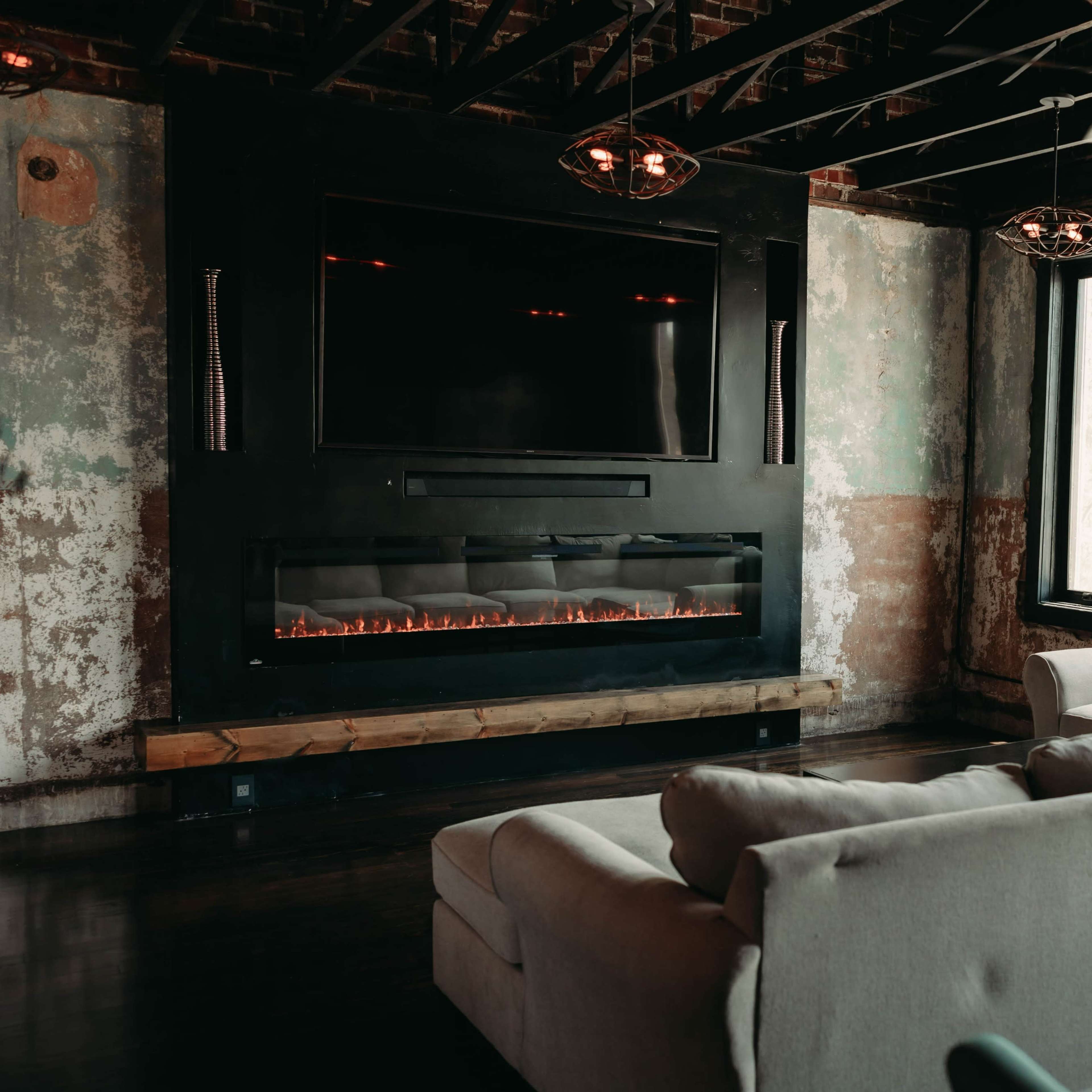 A modern living room features a large sectional sofa facing an electric fireplace mounted beneath a flat-screen TV, with large windows allowing natural light to enter the space.