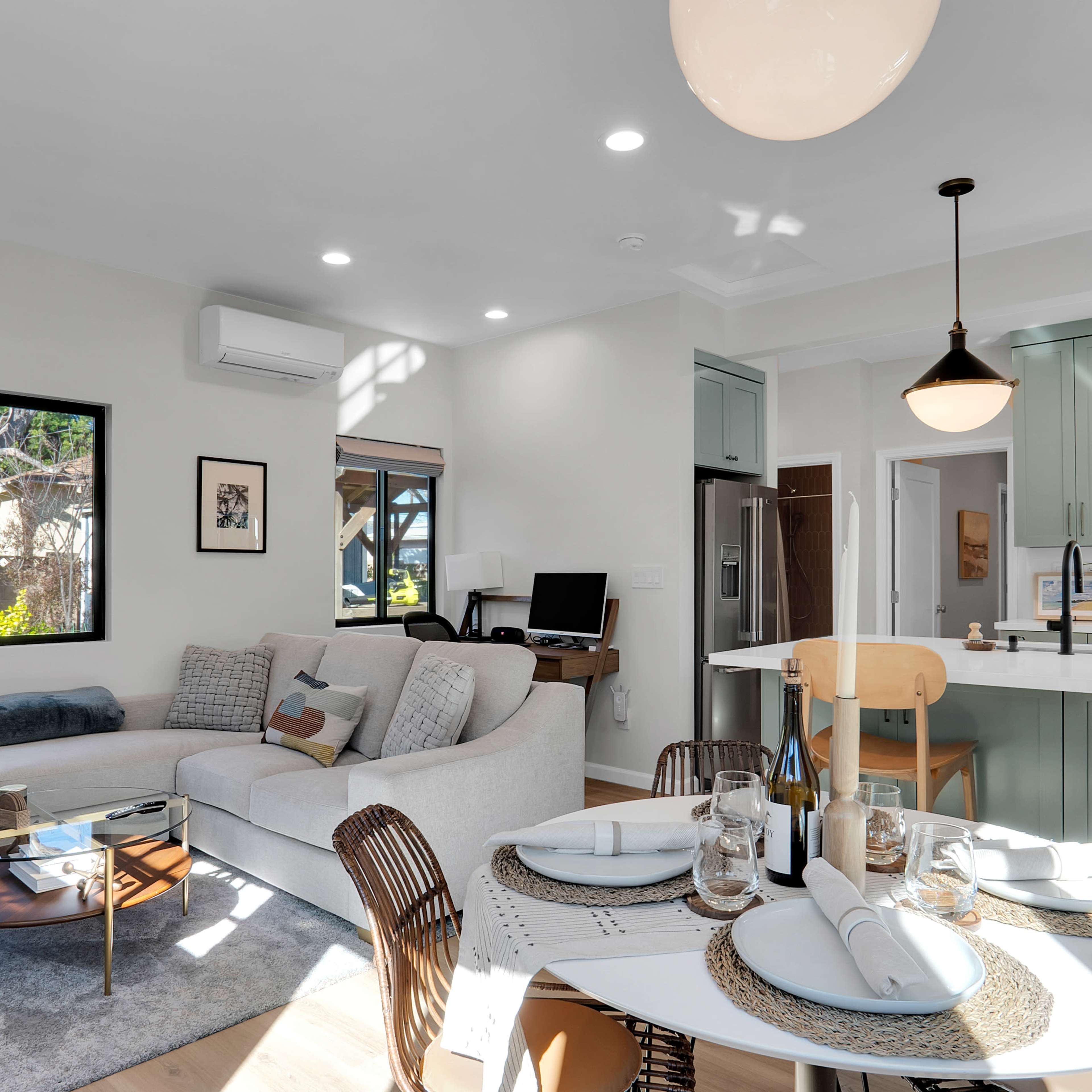 A modern living space features a light-colored couch, a dining table set for four, and a kitchen with green cabinetry.