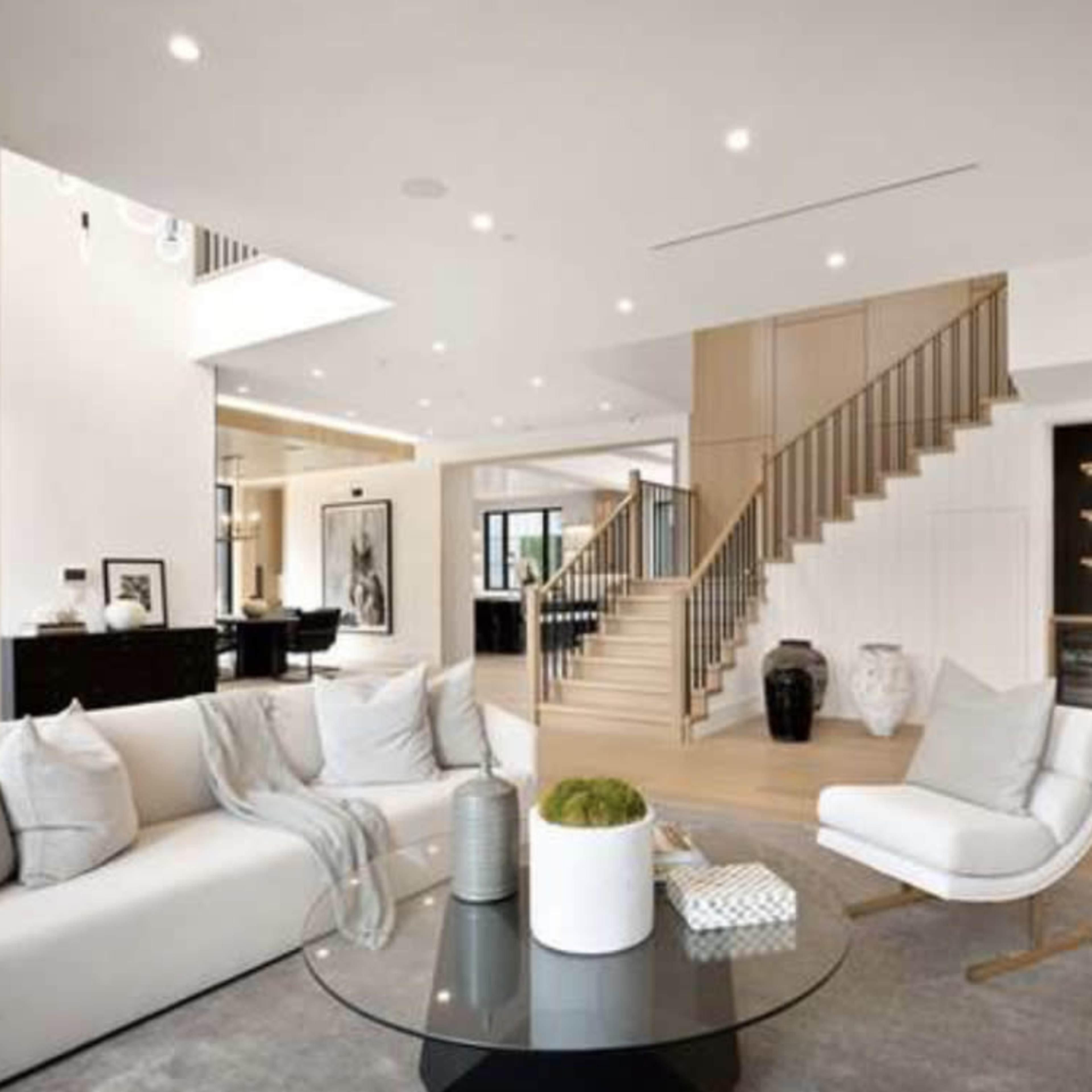 The room features a modern living space with a white sectional sofa, a circular glass table, and a wooden staircase leading to an upper level.
