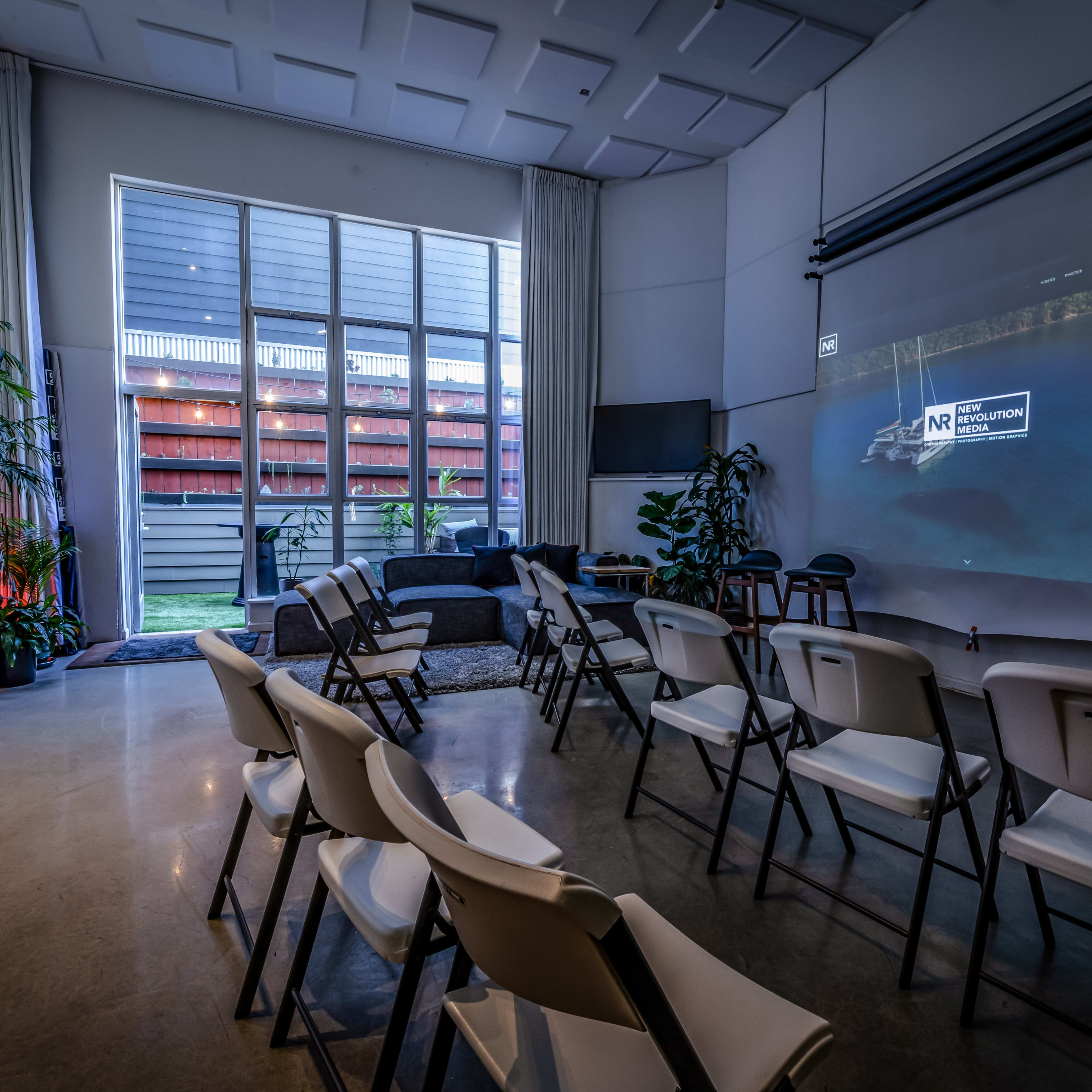 A modern room contains rows of white chairs facing a projection screen, with large windows revealing an outdoor space.