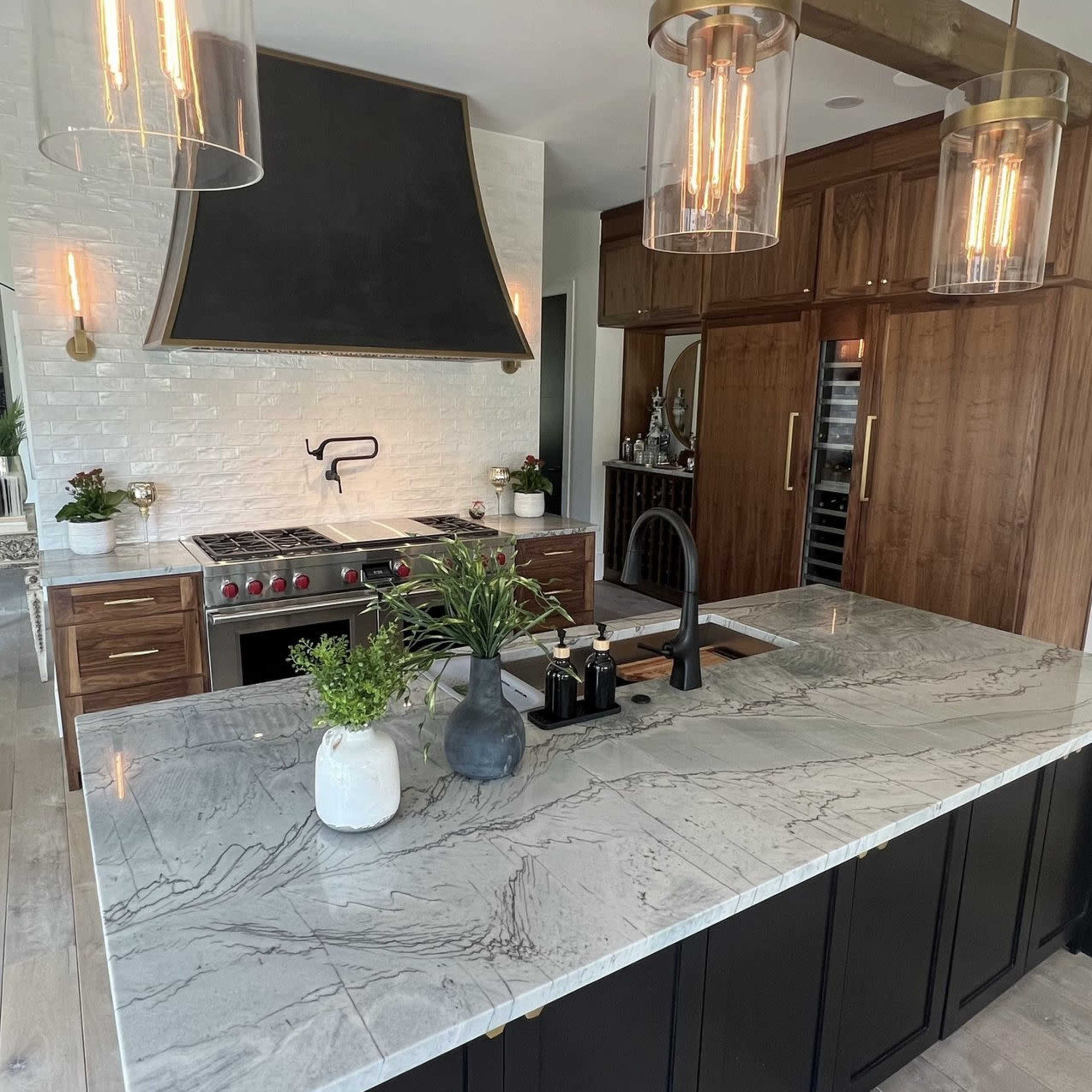 A modern kitchen featuring a large marble island, dark cabinetry, a decorative range hood, and stainless steel appliances.