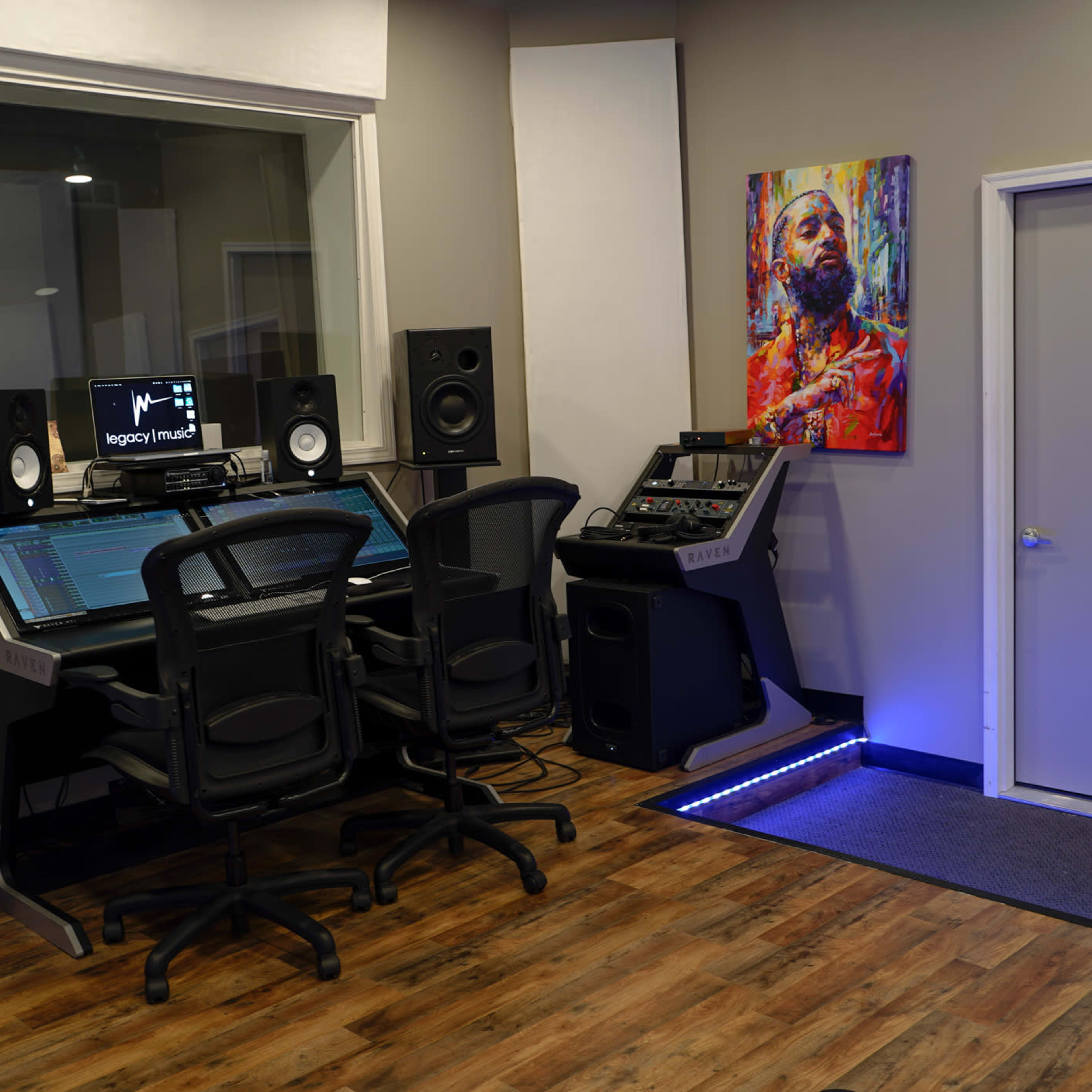 A music production studio with a control desk, two speakers, three chairs, and a colorful painting on the wall.