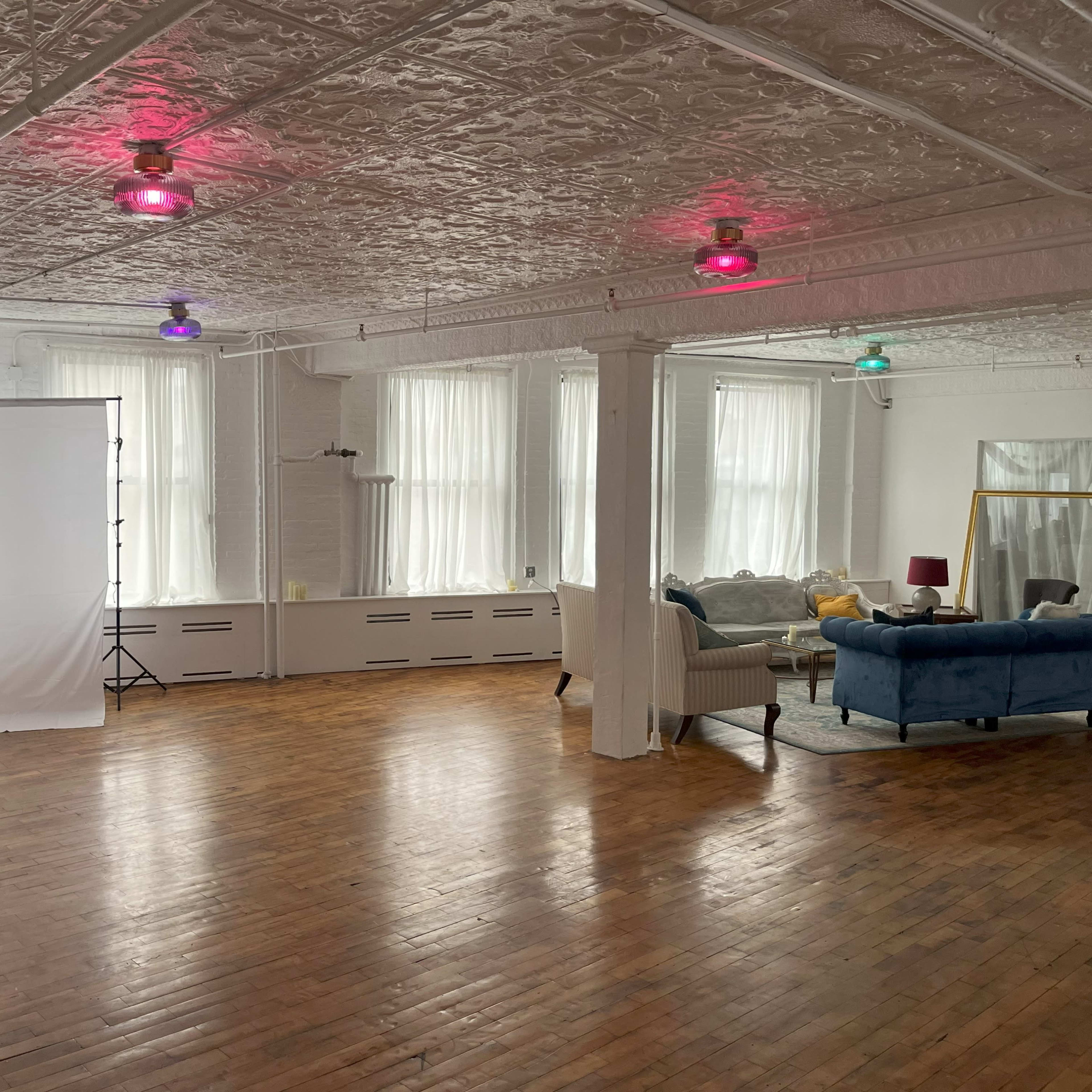 A spacious, light-filled studio with a white backdrop on one side and a furnished seating area featuring a blue couch and a large mirror on the other.