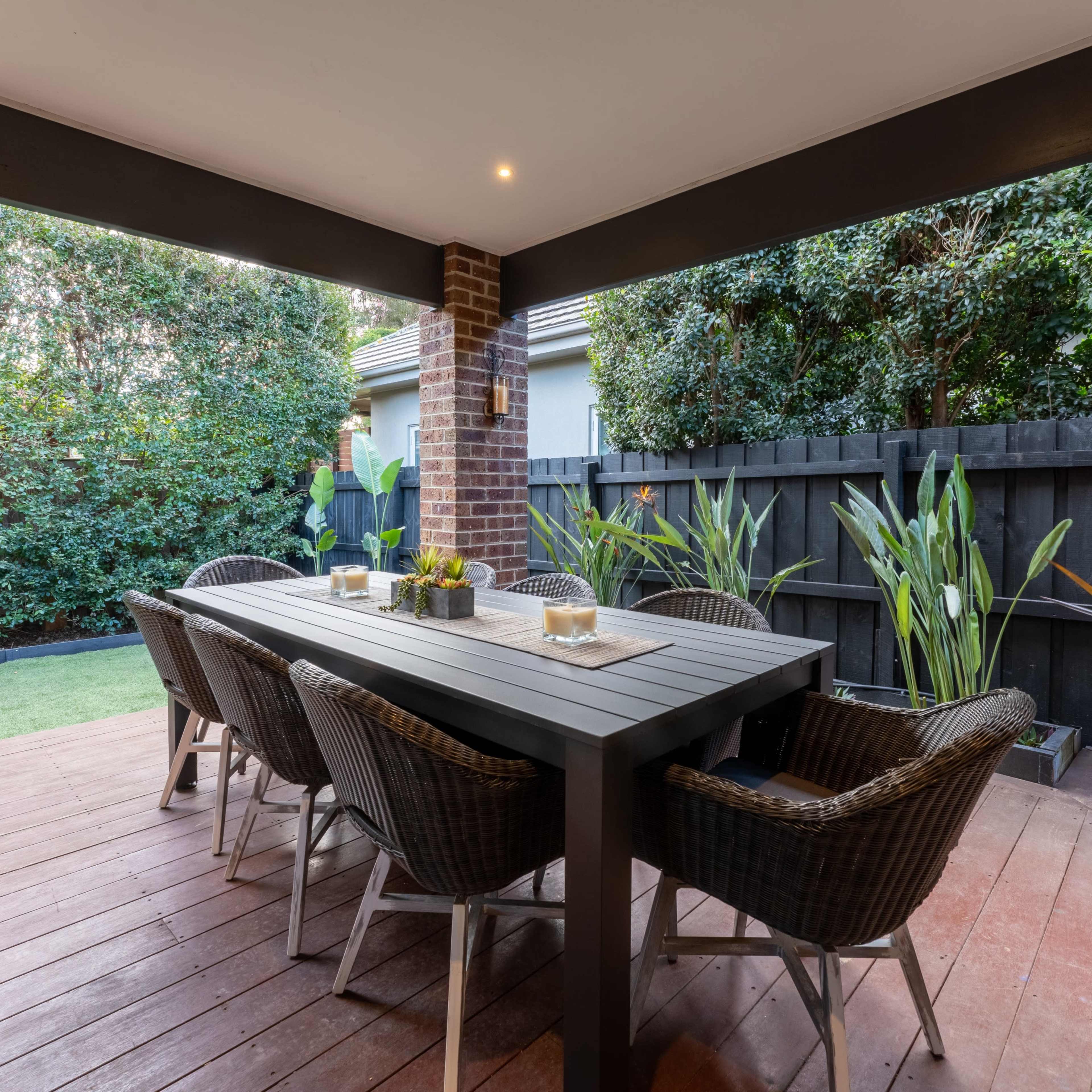 A covered outdoor dining area with a long table and six wicker chairs, surrounded by greenery and a dark wooden fence.